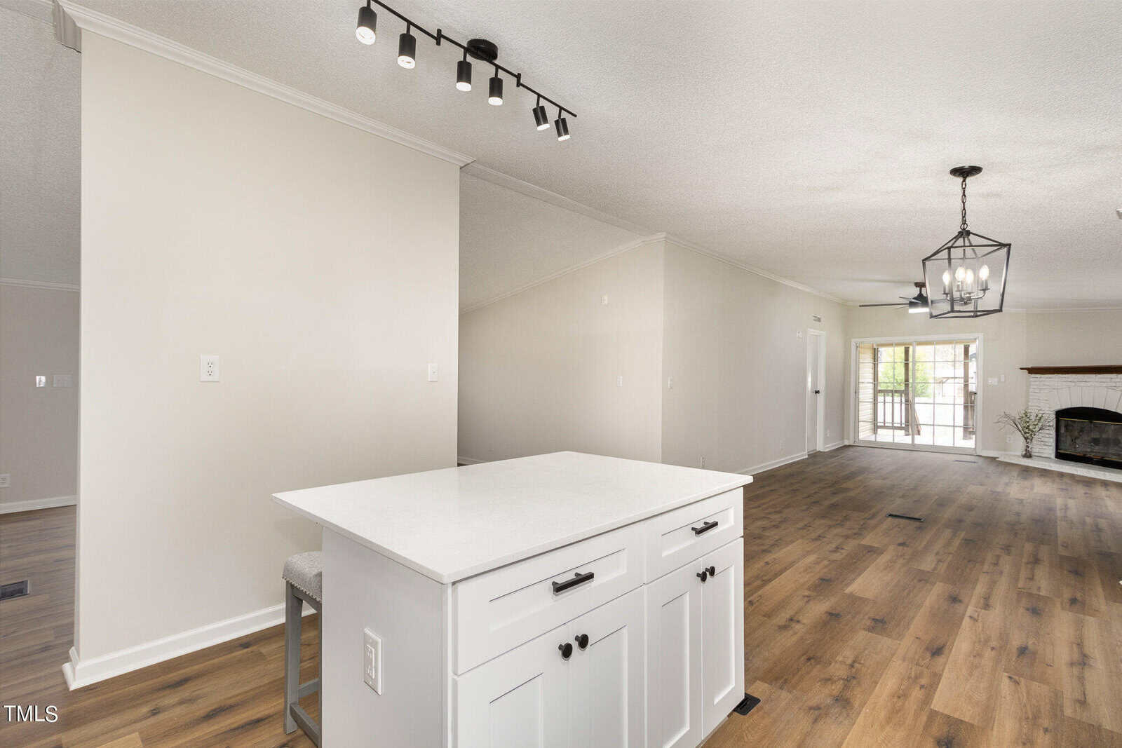 Spacious open-concept living area with a kitchen island featuring white cabinetry. The area has light wooden flooring, a track light on the ceiling, and a hanging lantern light fixture. A fireplace is visible in the background near large windows.