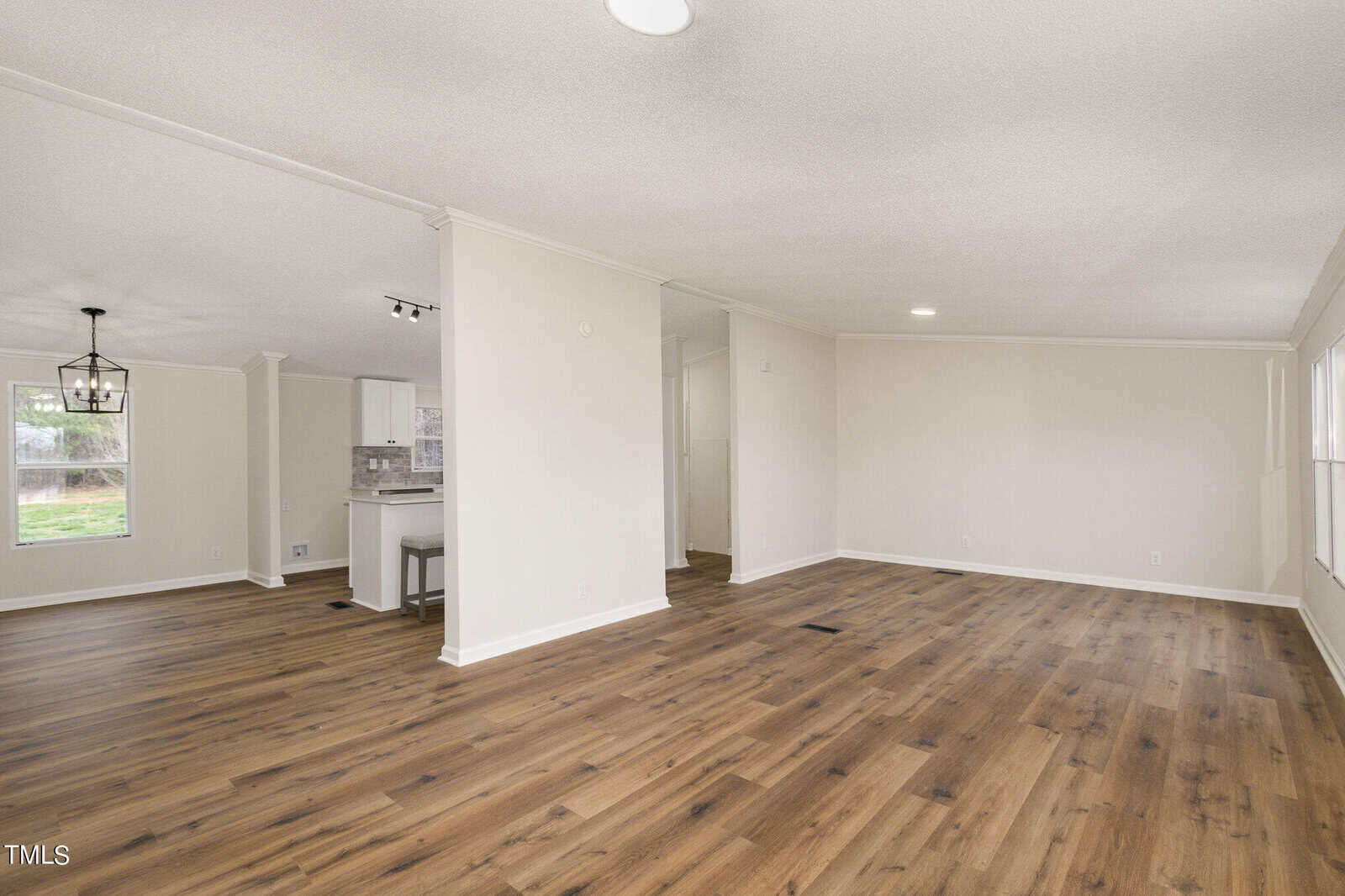 A spacious open-plan living area with light beige walls, recessed lighting, and wood laminate flooring. The kitchen, to the left, features white cabinets and a breakfast bar. A large window lets in natural light.