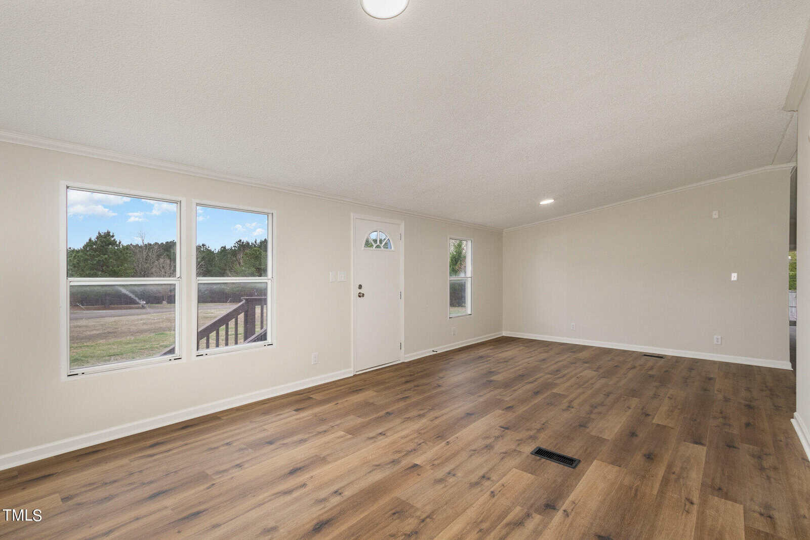 A spacious, empty living room with light-colored walls, a wooden floor, and large windows that let in natural light. A white door with a small window is visible, and trees can be seen outside.