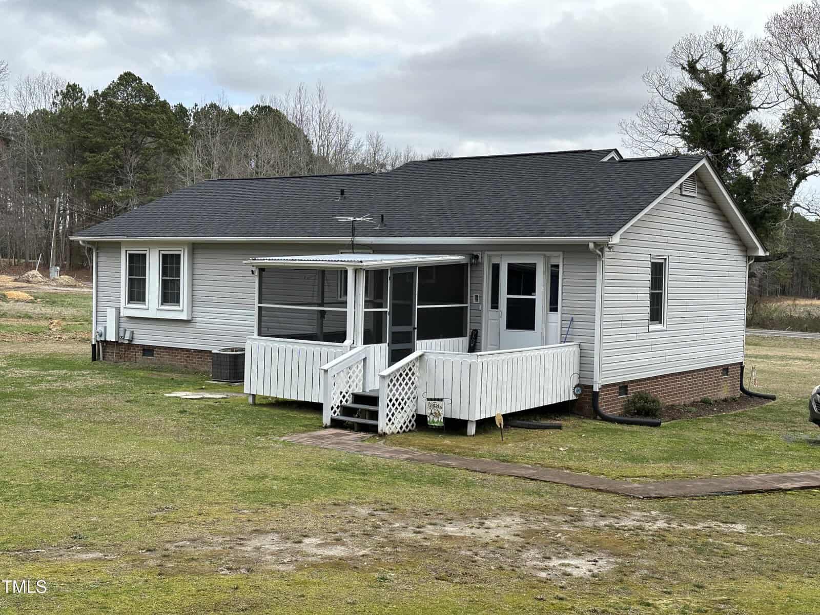 A single-story white house with a covered, screened porch, sloped roofing, and surrounded by a large, open grassy yard. a walkway leads up to the porch, and the sky is overcast.