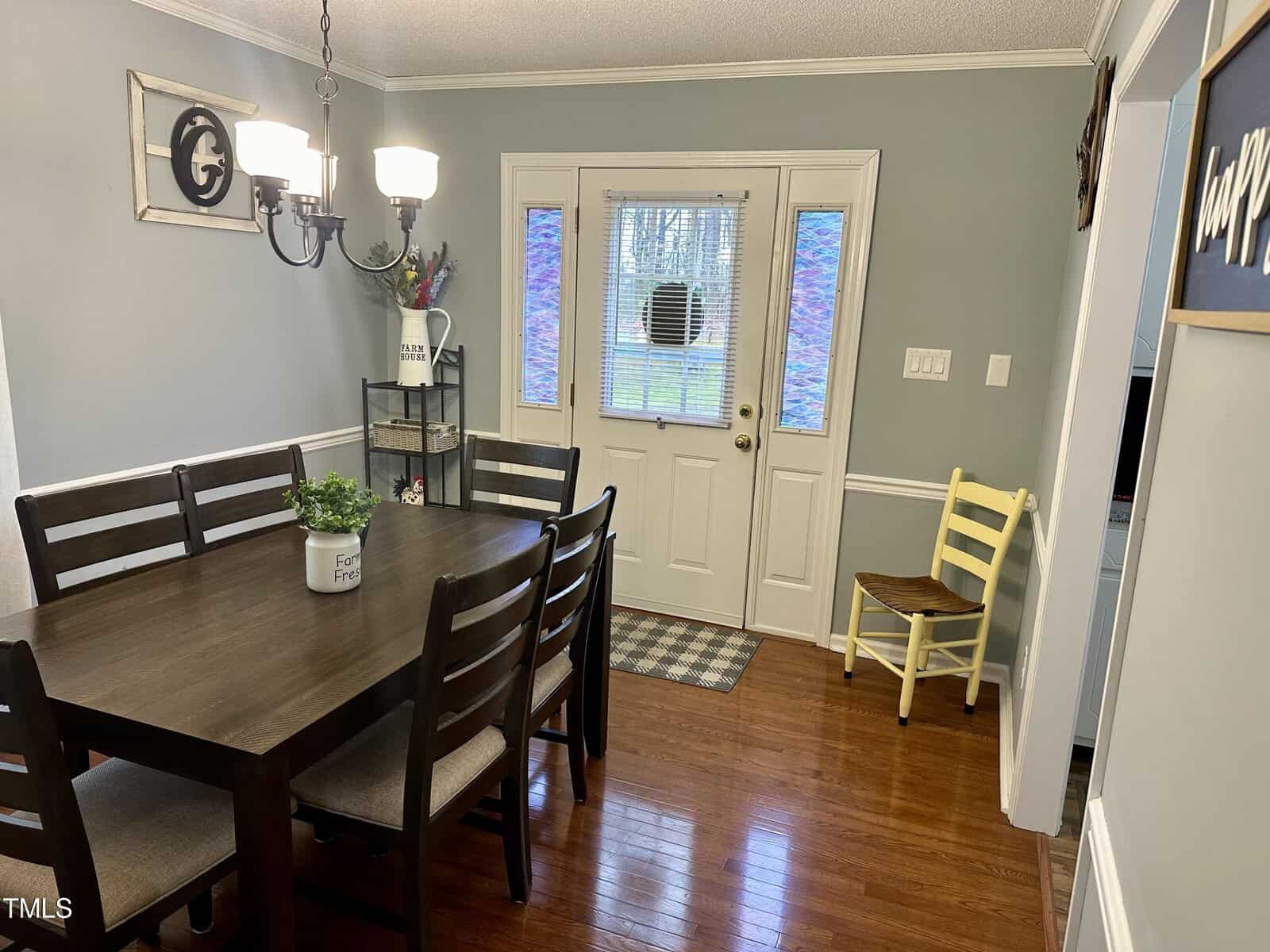 A dining room featuring a wooden table with six chairs, gray walls, a white door with a window, and a yellow chair beside a small side table. decor includes a wall clock and framed art.