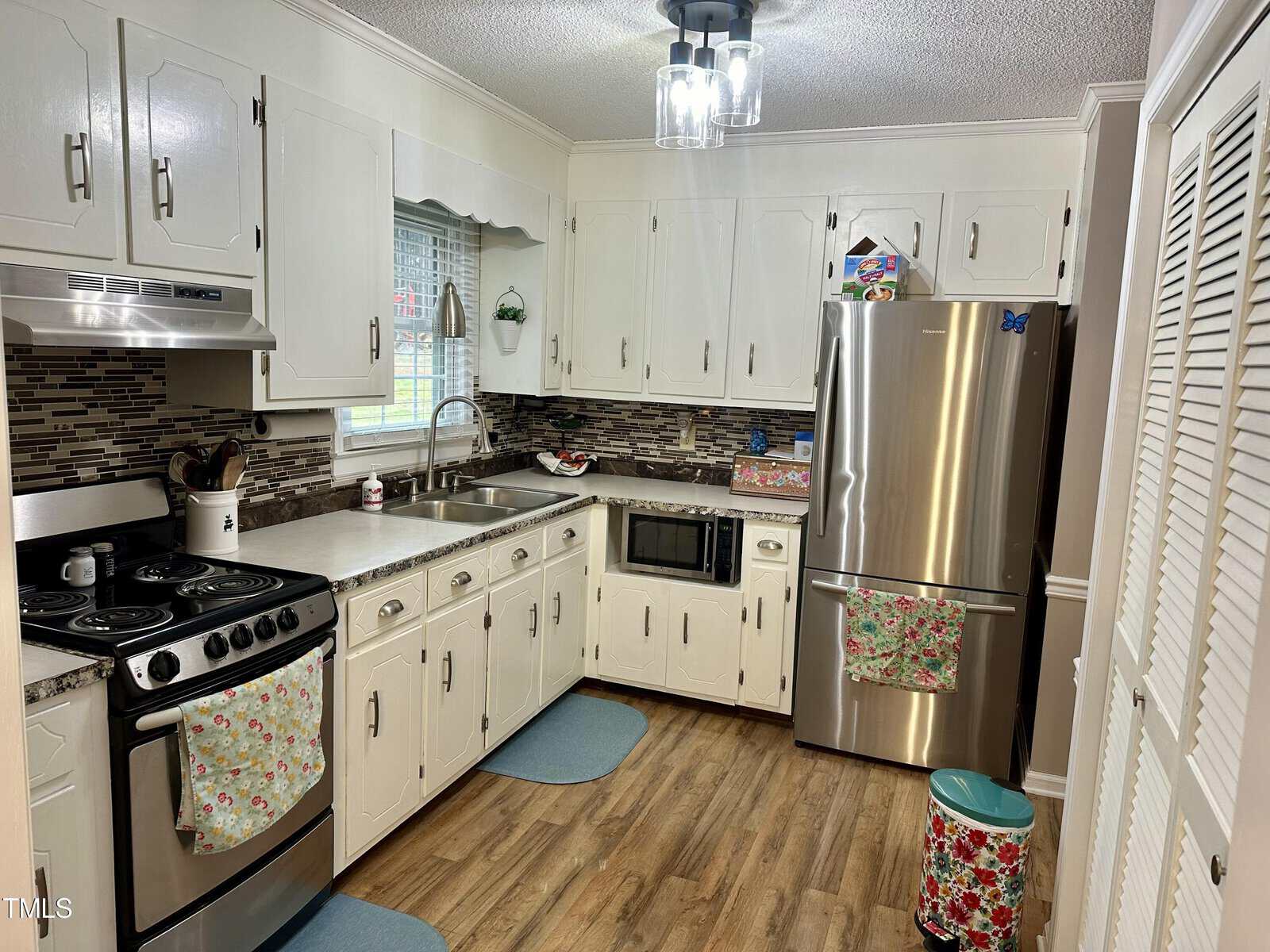 A cozy kitchen with white cabinets, brick backsplash, stainless steel appliances, and hardwood flooring. it features a window above the sink and bright overhead lighting.