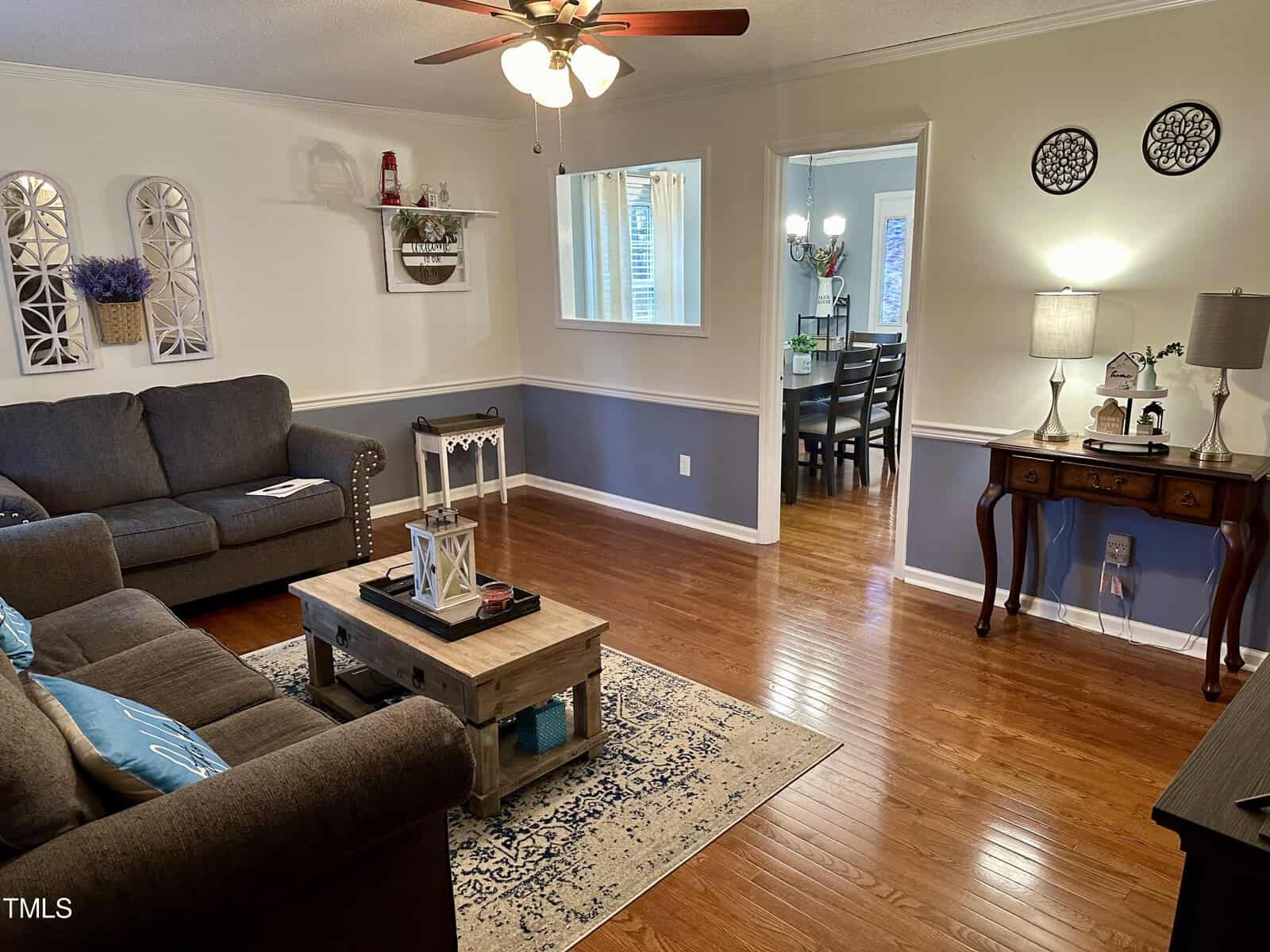 Interior view of a living room with dark hardwood floors, two grey sofas, and a small coffee table. a decorative rug is placed in the center. there's a view into a dining area with blue walls.