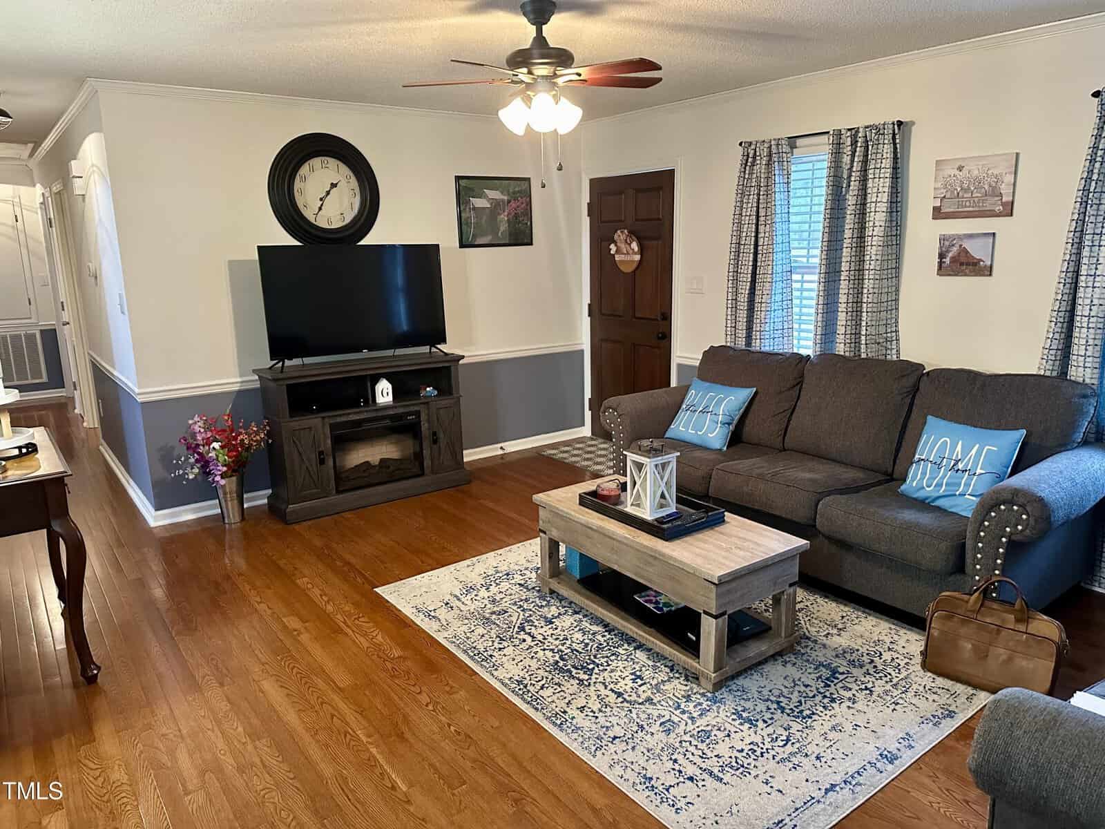A cozy living room featuring a dark gray sectional sofa, a wooden coffee table, and a tv on a media stand. the room has hardwood floors, white curtains, and decorative elements like a large wall clock.