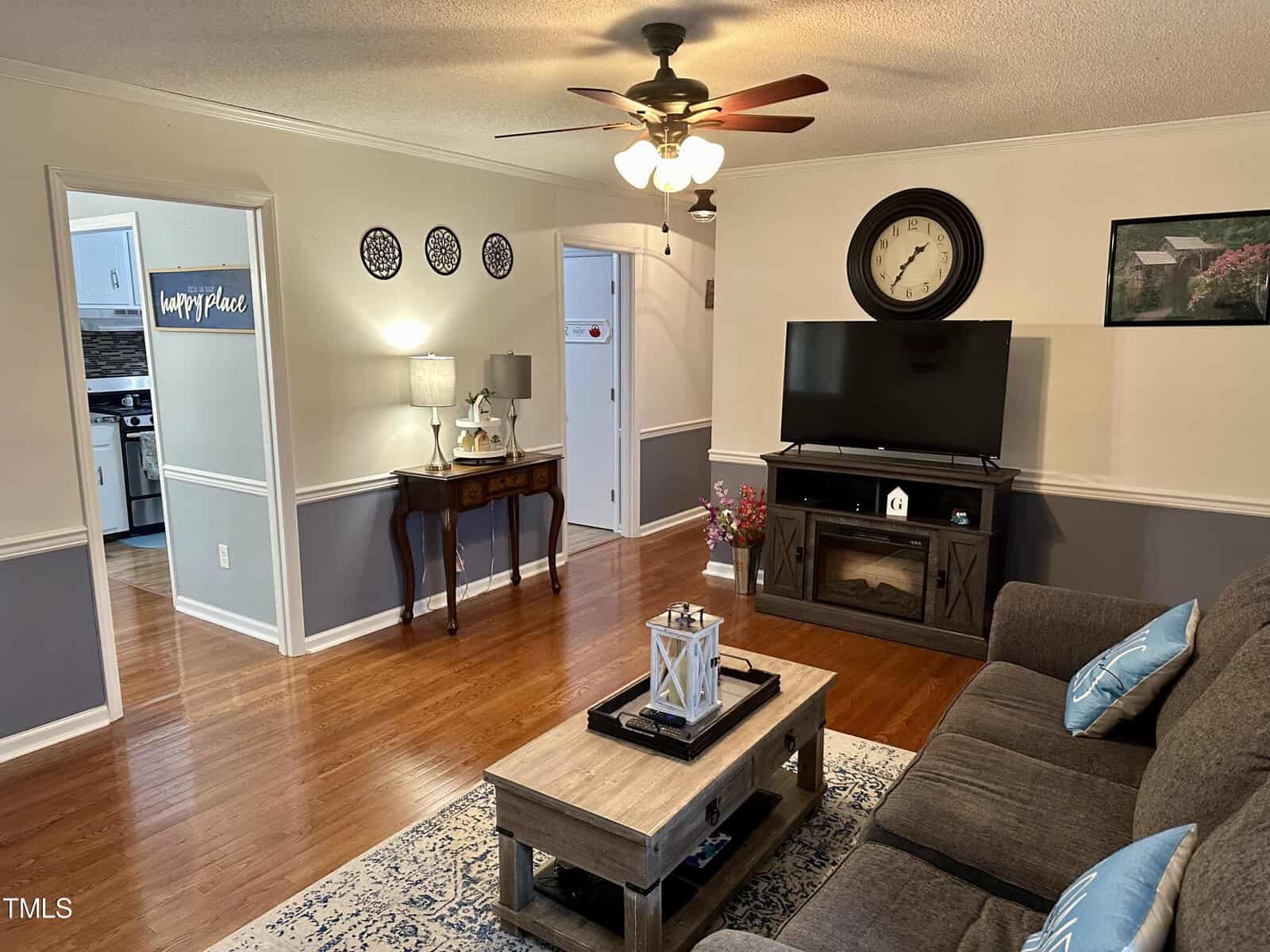 Living room with a gray sofa, wooden coffee table, and tv stand. features a ceiling fan, decorative plates on the wall, and a doorway leading to a kitchen. bright and neatly arranged.