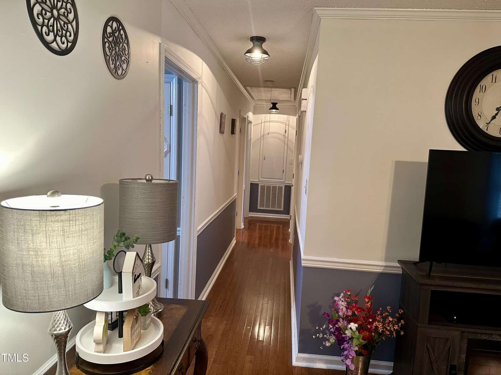 An interior view of a narrow hallway in a house, showing a wooden floor, white walls, and a series of doors on one side. decorative items include wall art, lamps on a console table, and a large clock.