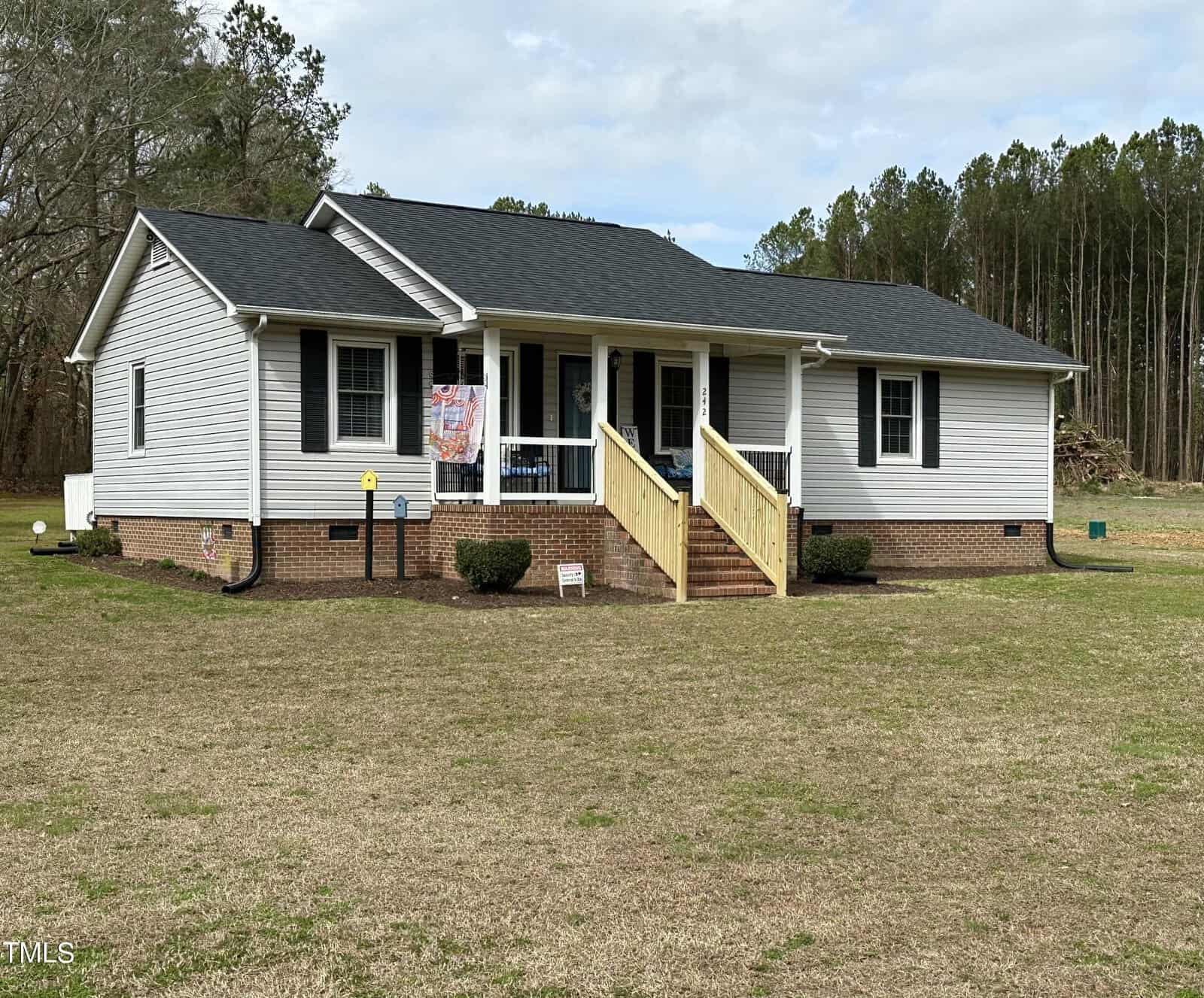 A one-story white house with a gabled roof, front porch with wooden steps, and several windows, set on a large lawn bordered by a forest at 242 Edwards Road, Henderson NC. A