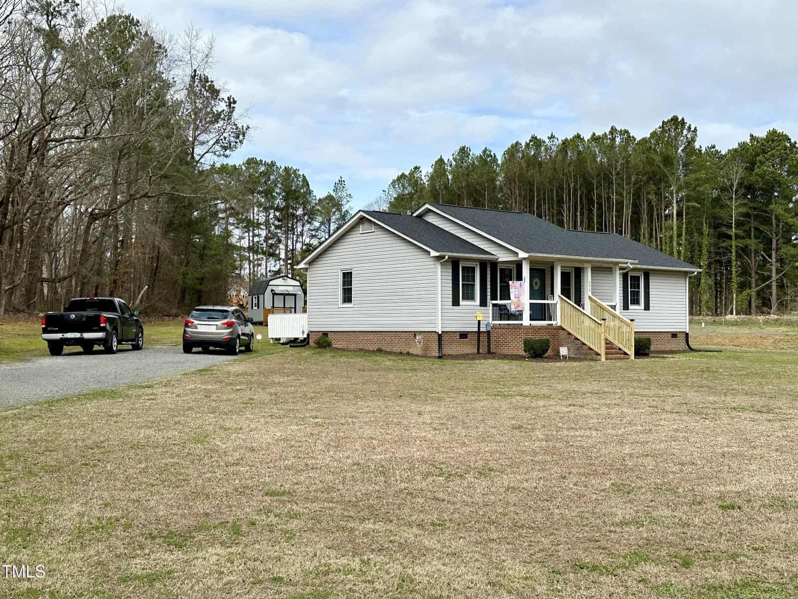 A one-story white house with a gabled roof surrounded by a grassy lawn and a backdrop of tall trees. in the driveway, there are three vehicles including an suv and a pickup truck.