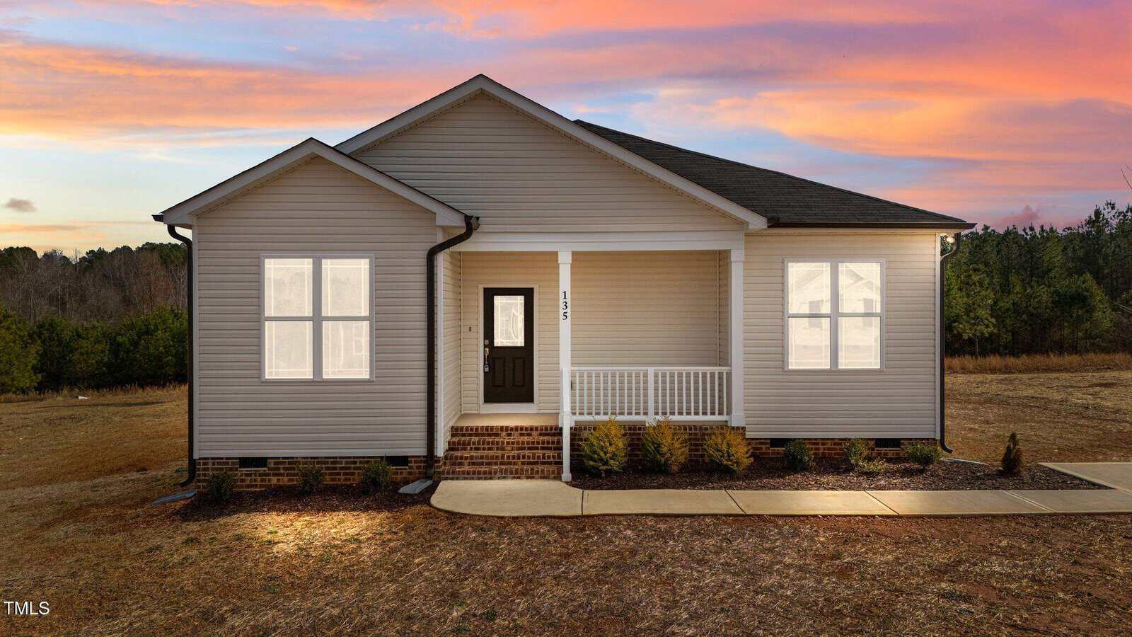 A single-story beige house with white trim and a dark roof, front porch, brick steps, and small shrubs in front, set against a sunset sky with trees in the background.