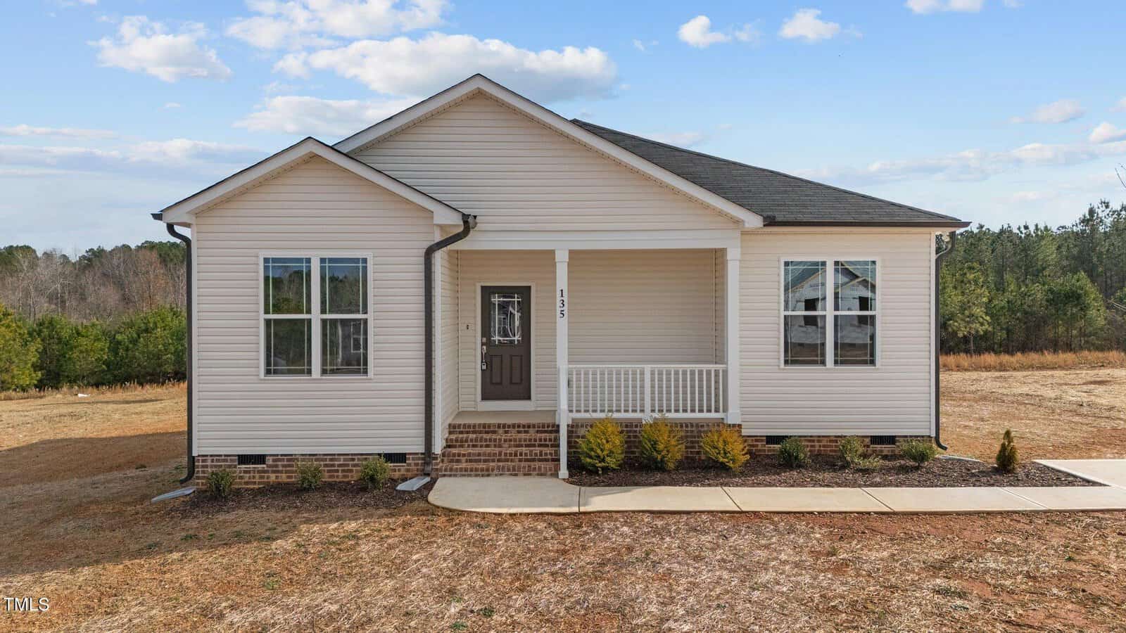 Single-story beige house with a covered front porch, dark roof, and small shrubs along the front. The home sits on a large, dry lot with scattered trees and blue sky in the background.