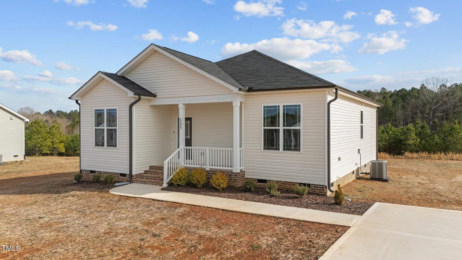 A small, single-story white house with black roof shingles, a front porch with steps, and large windows sits on a dry, patchy yard with minimal landscaping and a concrete driveway under a partly cloudy sky.
