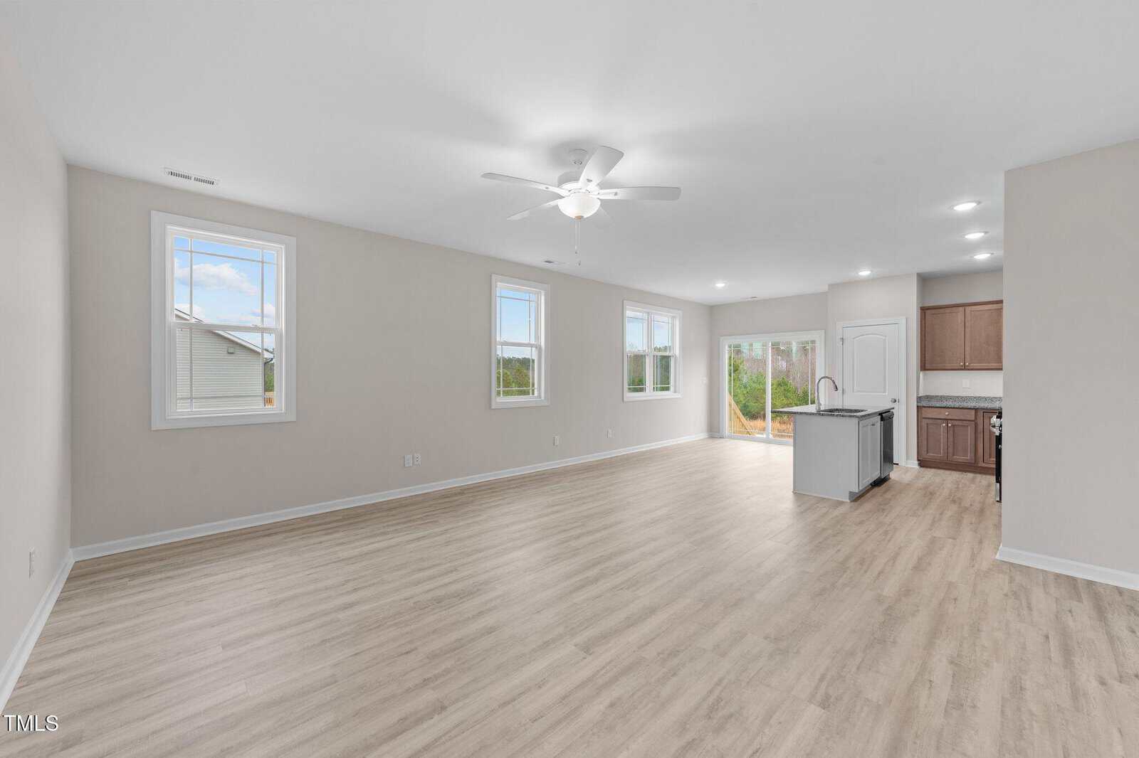 Spacious, empty open-concept living area with light wood flooring, beige walls, ceiling fan, large windows, and a modern kitchen with brown cabinets and an island, next to glass doors leading outside.