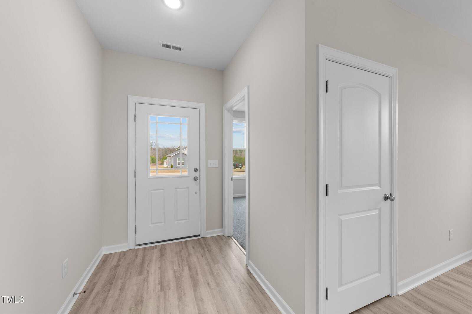 A bright entryway with light wood flooring, beige walls, a white front door with glass panes, a white six-panel door to the right, and a room with blue carpet visible through an open doorway.