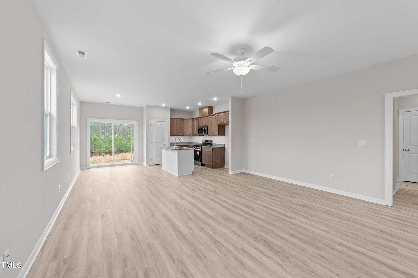 Spacious open-plan living area with light wood flooring, a ceiling fan, large windows, and a kitchen with wood cabinets, stainless steel appliances, and a center island. Sliding glass door opens to a wooded backyard view.