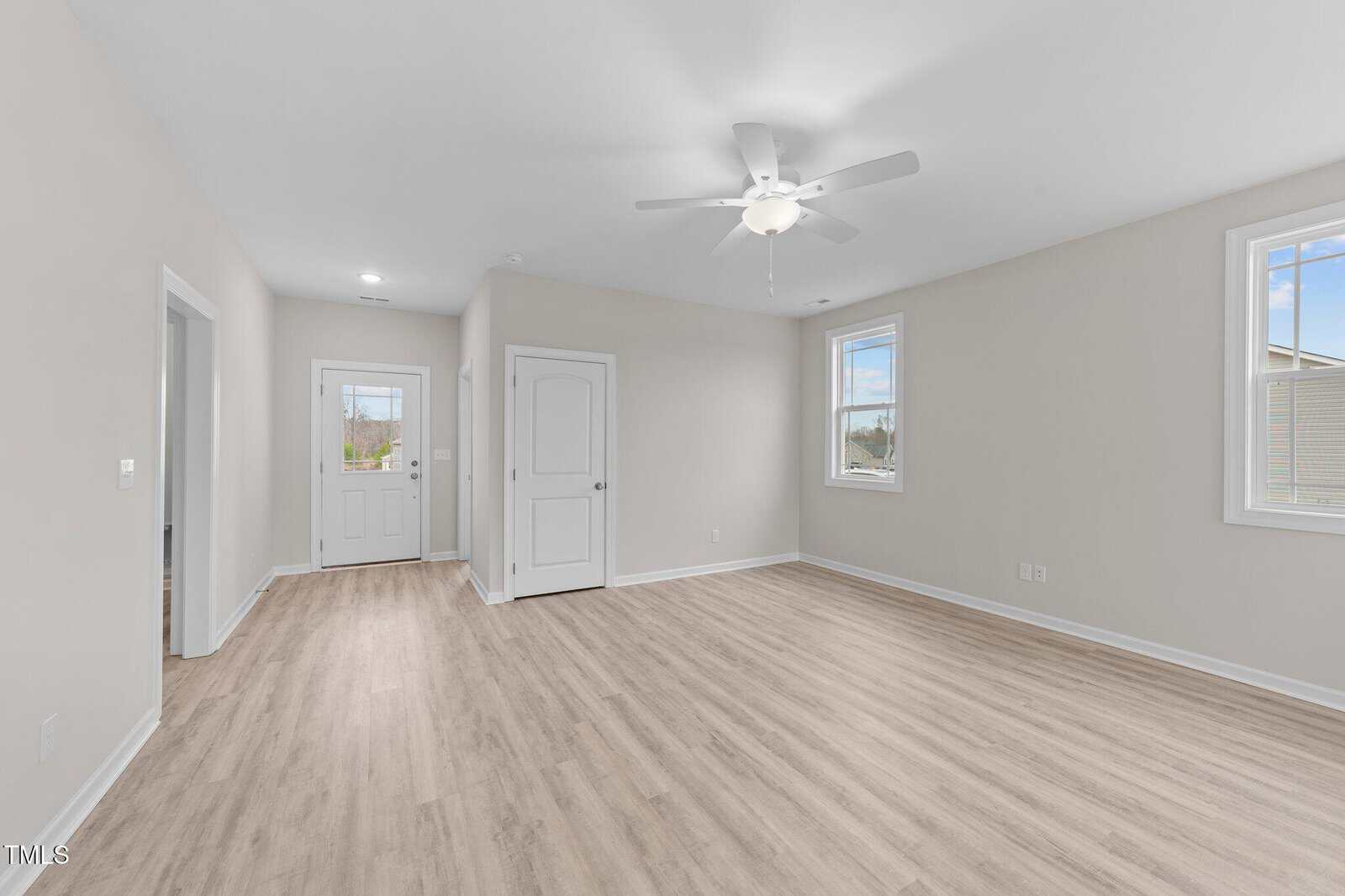 A bright, empty room with light wood flooring, neutral walls, two windows, a white ceiling fan, and three doors. Natural light streams in, creating a clean, modern, and spacious atmosphere.