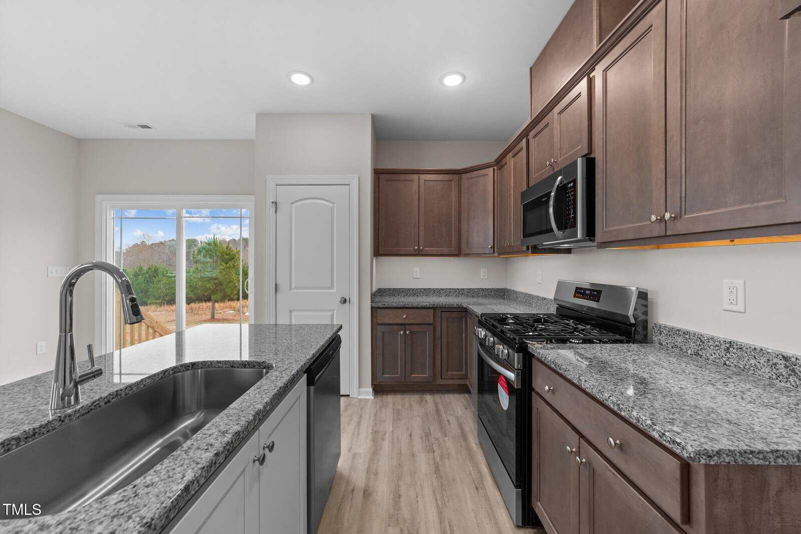 Modern kitchen with brown cabinets, granite countertops, stainless steel appliances, gas stove, built-in microwave, and a sink in the island. Sliding glass doors lead to a backyard with trees. Light wood flooring throughout.