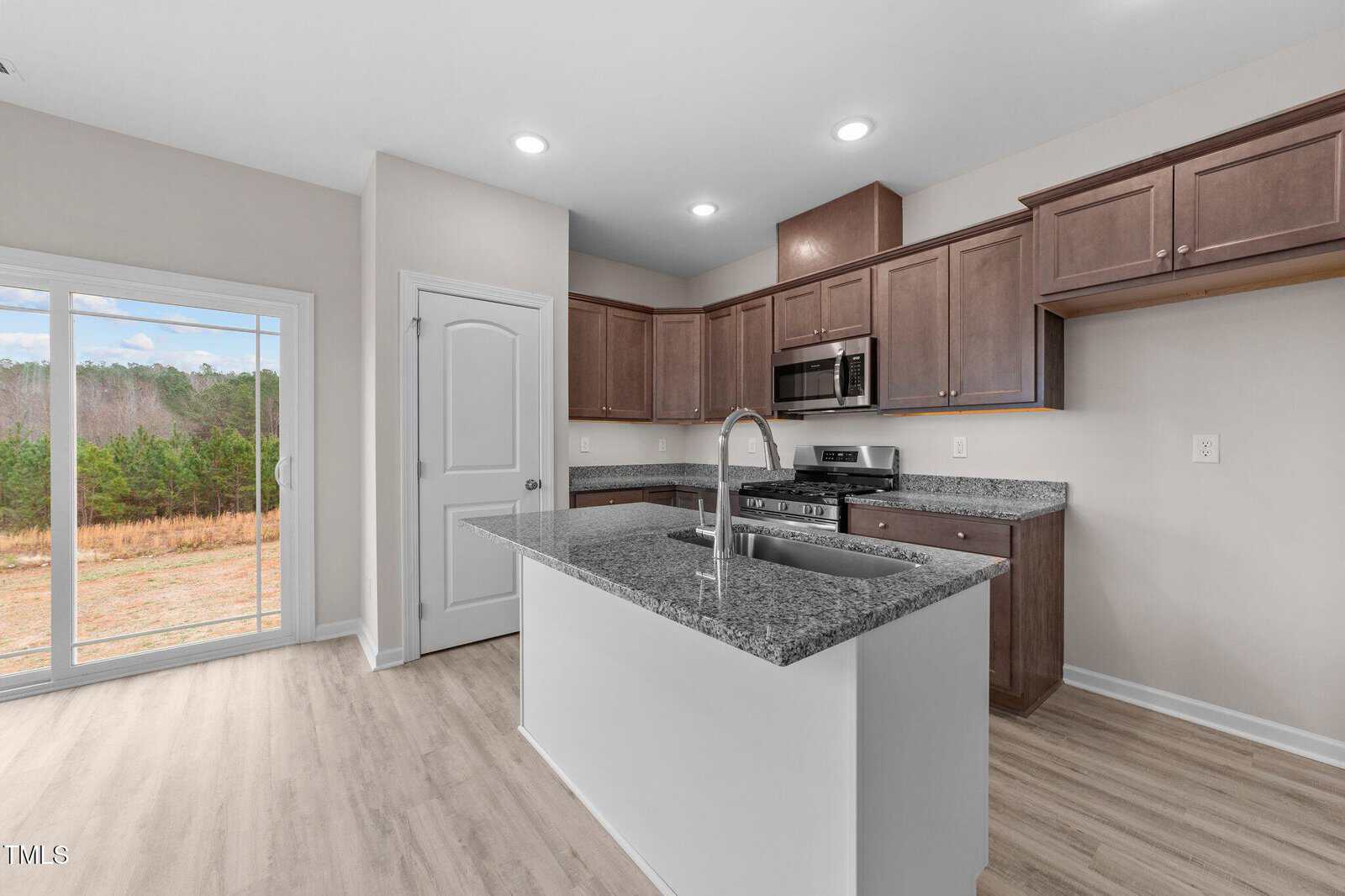 Modern kitchen with wood cabinets, granite countertops, stainless steel appliances, and a center island. There is a sliding glass door leading to an outdoor view of trees and a pantry door on the left wall.