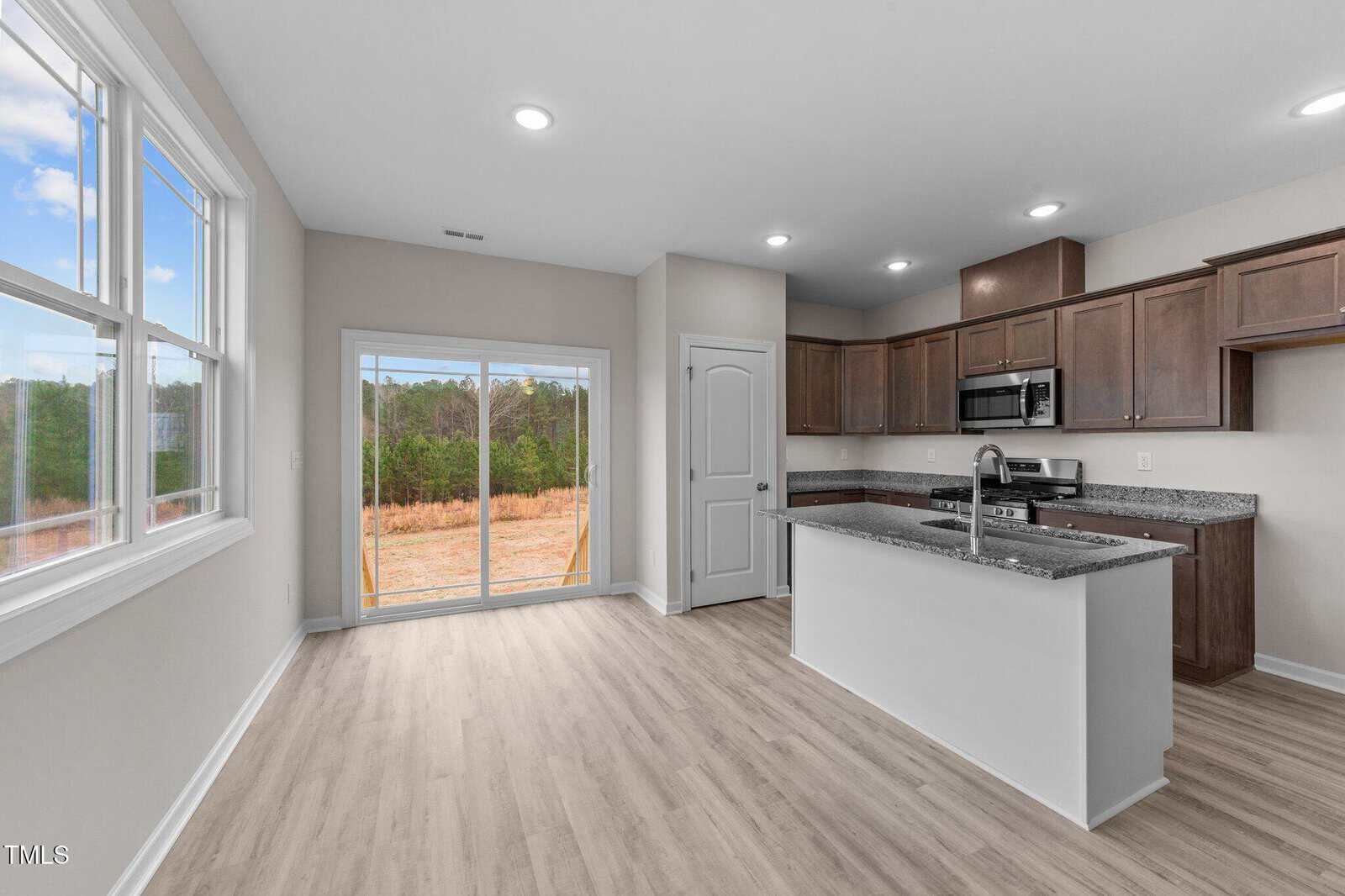 Modern kitchen and dining area with light wood floors, dark brown cabinets, granite countertops, stainless steel appliances, large windows, and sliding glass doors leading to a grassy backyard with trees.