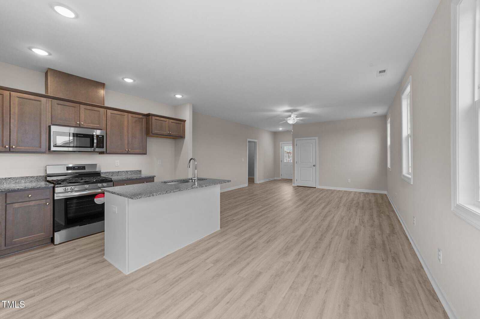 Open-concept kitchen and living area with light wood flooring, brown cabinets, granite countertops, stainless steel appliances, and neutral-colored walls. Several windows and recessed lighting create a bright, spacious atmosphere.