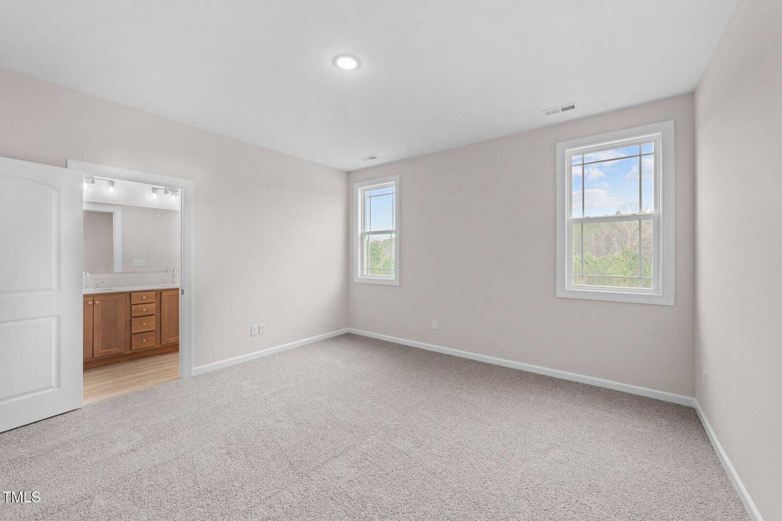 Empty bedroom with light beige walls, two windows, and carpeted floor. An open door leads to a bathroom with a double vanity and large mirror. The room is well-lit with a ceiling light.