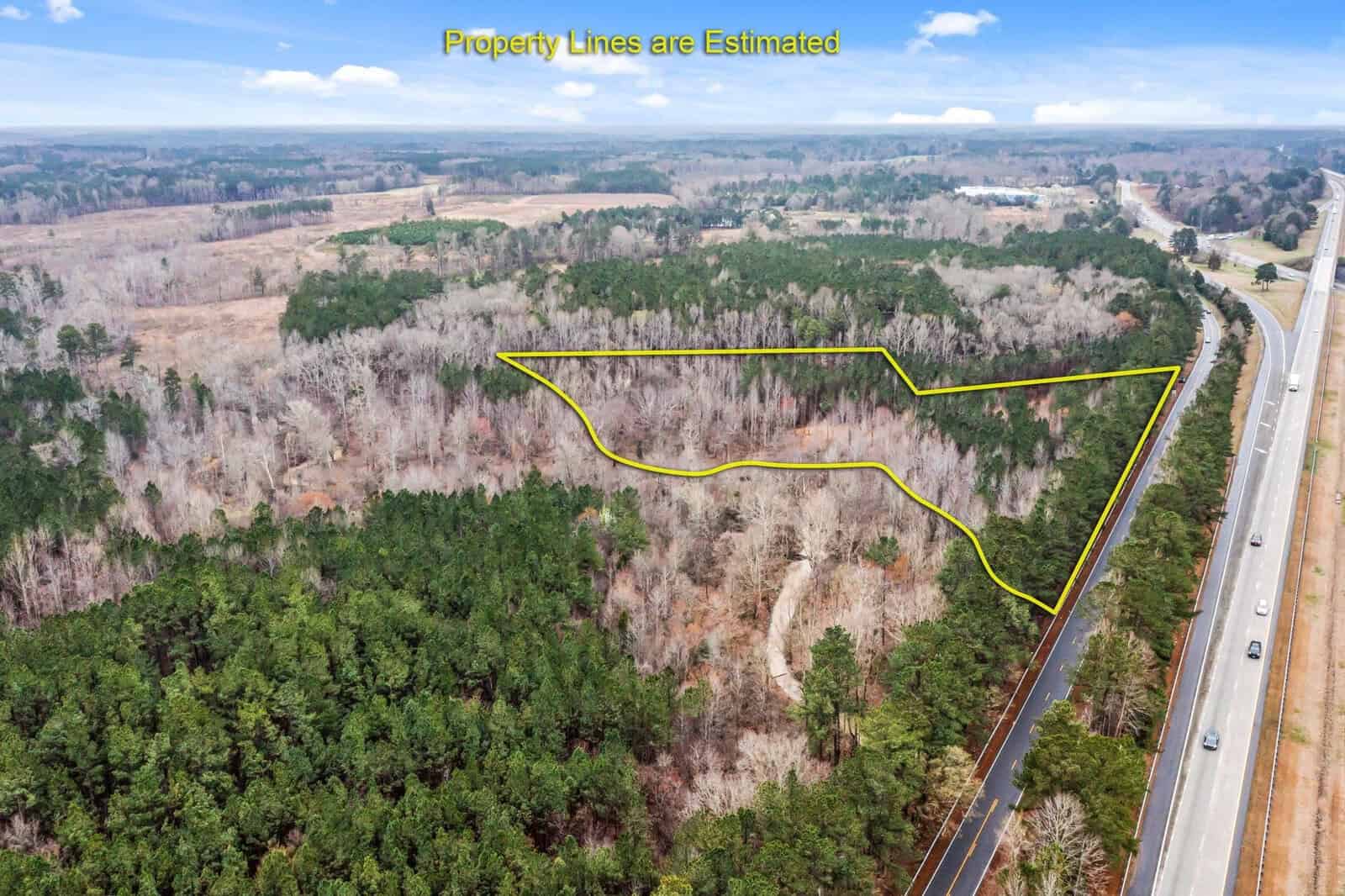 Aerial view of 3100 Knotts Grove Road, Oxford, with an estimated property boundary outlined in yellow. A road runs along the right side with a few vehicles visible. The landscape consists of dense trees and distant fields under a partly cloudy sky.