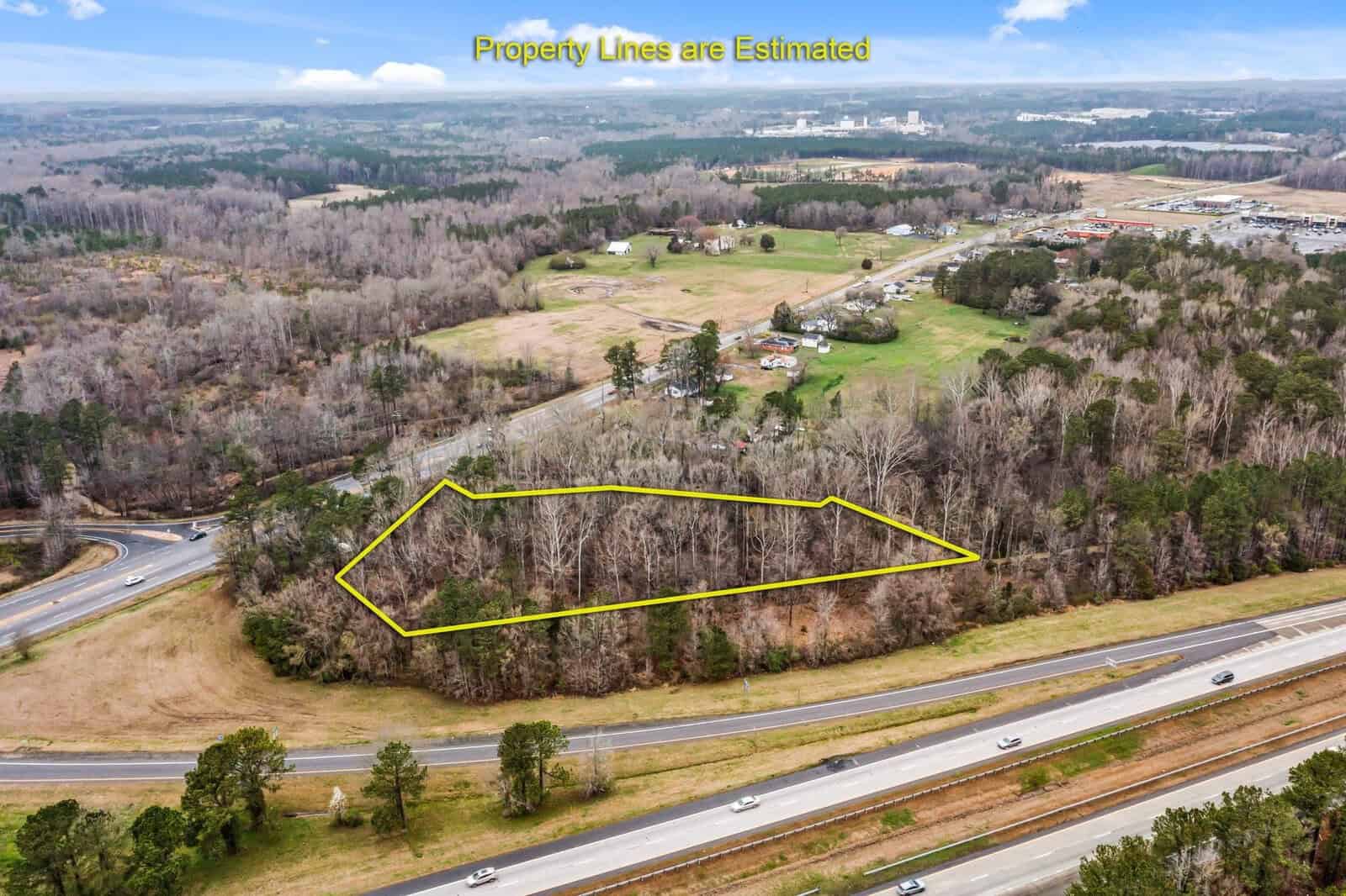Aerial view of a wooded area with an estimated property boundary outlined in yellow. Various roadways and highways surround the plot of land. Buildings and open fields are visible in the background under a partly cloudy sky.