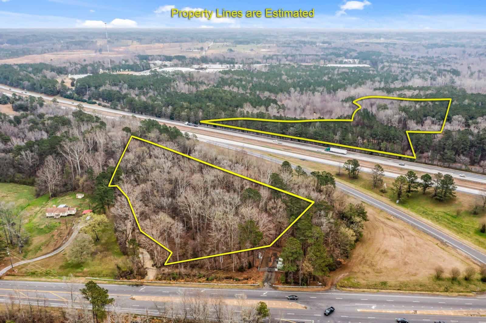 Aerial view of a landscape with two wooded areas outlined in yellow, indicating estimated property lines. A highway runs between the areas, with surrounding greenery and trees. A few buildings are visible in the lower left corner.