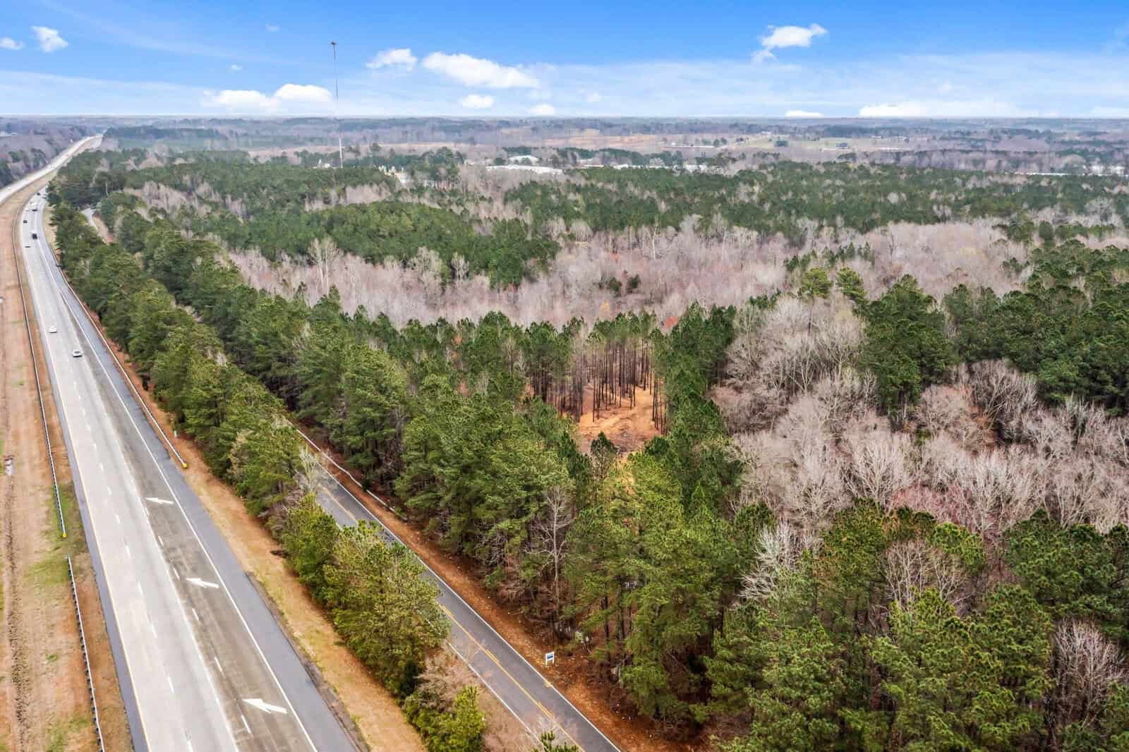 Aerial view of a highway running parallel to a dense forested area. The road is relatively empty with a few visible cars. The landscape features a mix of green coniferous trees and leafless deciduous trees under a blue sky with scattered clouds.