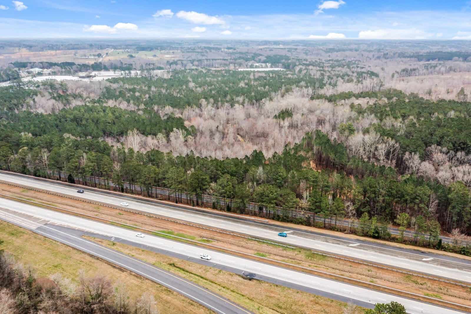 Aerial view of a highway with light traffic, surrounded by dense forest on a clear day. The landscape includes vast stretches of trees and a blue sky with scattered clouds.