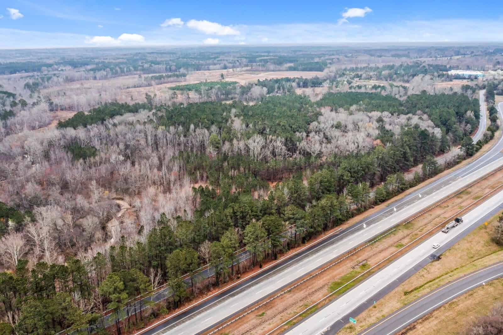 Aerial view of a highway running through a forested area. The road is divided into two lanes, with a few vehicles visible. Surrounding the highway are dense clusters of trees, with a mix of evergreen and leafless branches, under a blue sky with clouds.