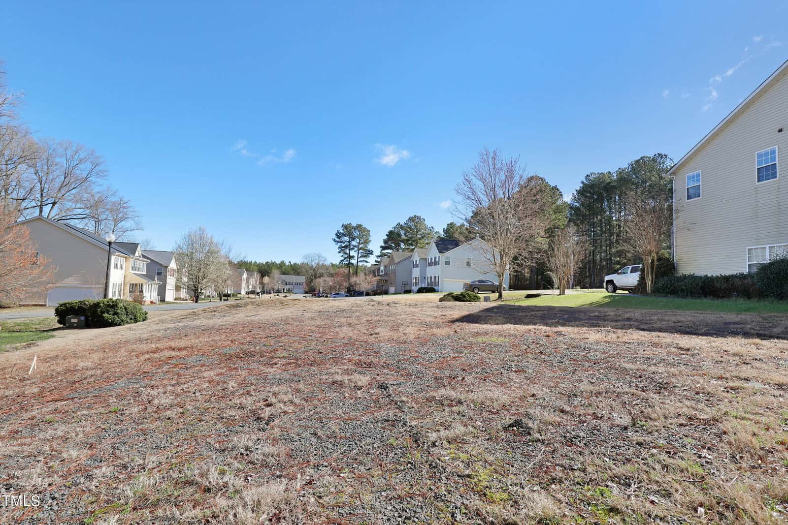 A vacant, grassy lot in a suburban neighborhood, bordered by houses, with a clear blue sky above. Trees are scattered in the background, and a white vehicle is parked along the street.