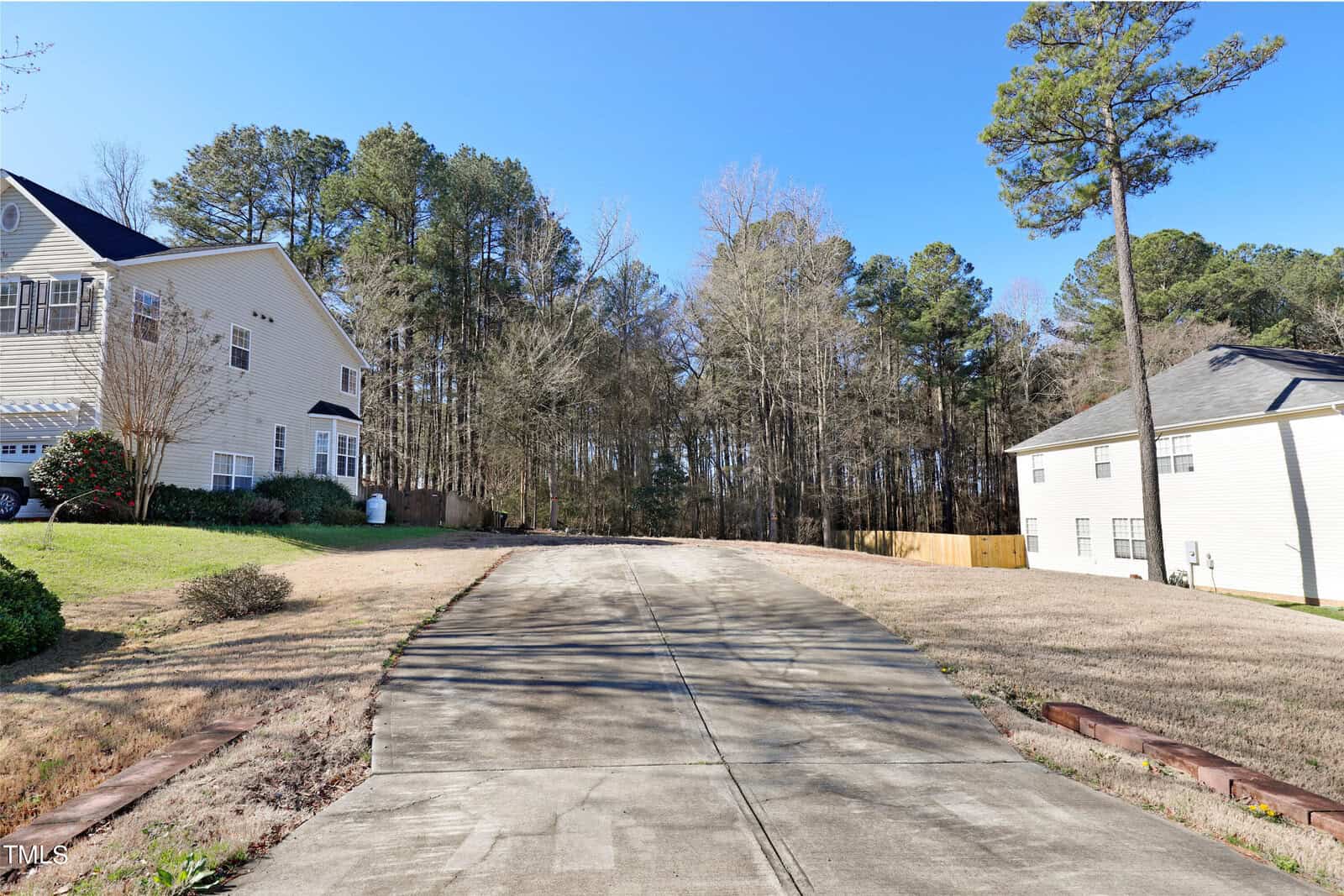 A long, empty concrete driveway leads to a wooded area lined with tall trees. On either side are two-story houses with light-colored exteriors and manicured lawns, under a clear blue sky.