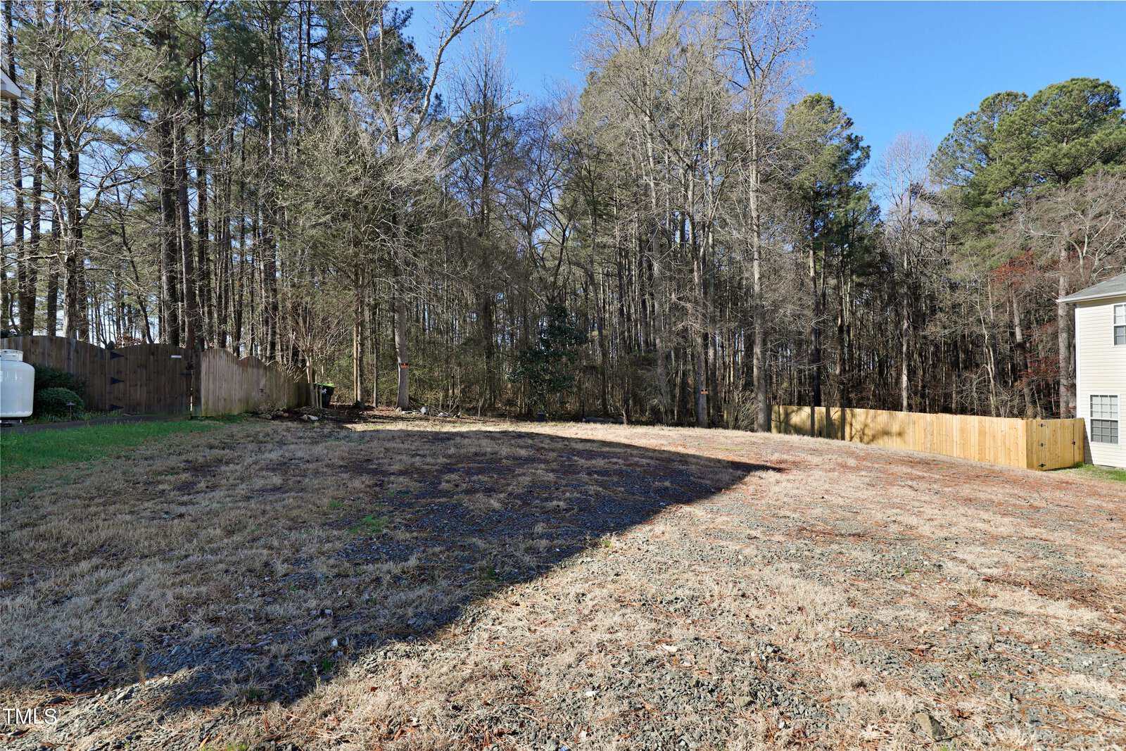 A sloped grassy yard with patches of dirt, bordered by a wooden fence on the left and right. A propane tank sits on the left side. Dense trees fill the background under a clear blue sky.