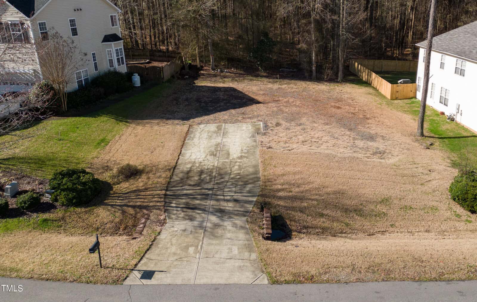 A vacant plot of land at 1603 Irving Place features a concrete driveway leading to it, nestled between two houses. It's bordered by a wooden fence on one side and flanked by a line of trees at the back. The grass is patchy, displaying both green and brown areas, embodying Creedmoor's rustic charm.