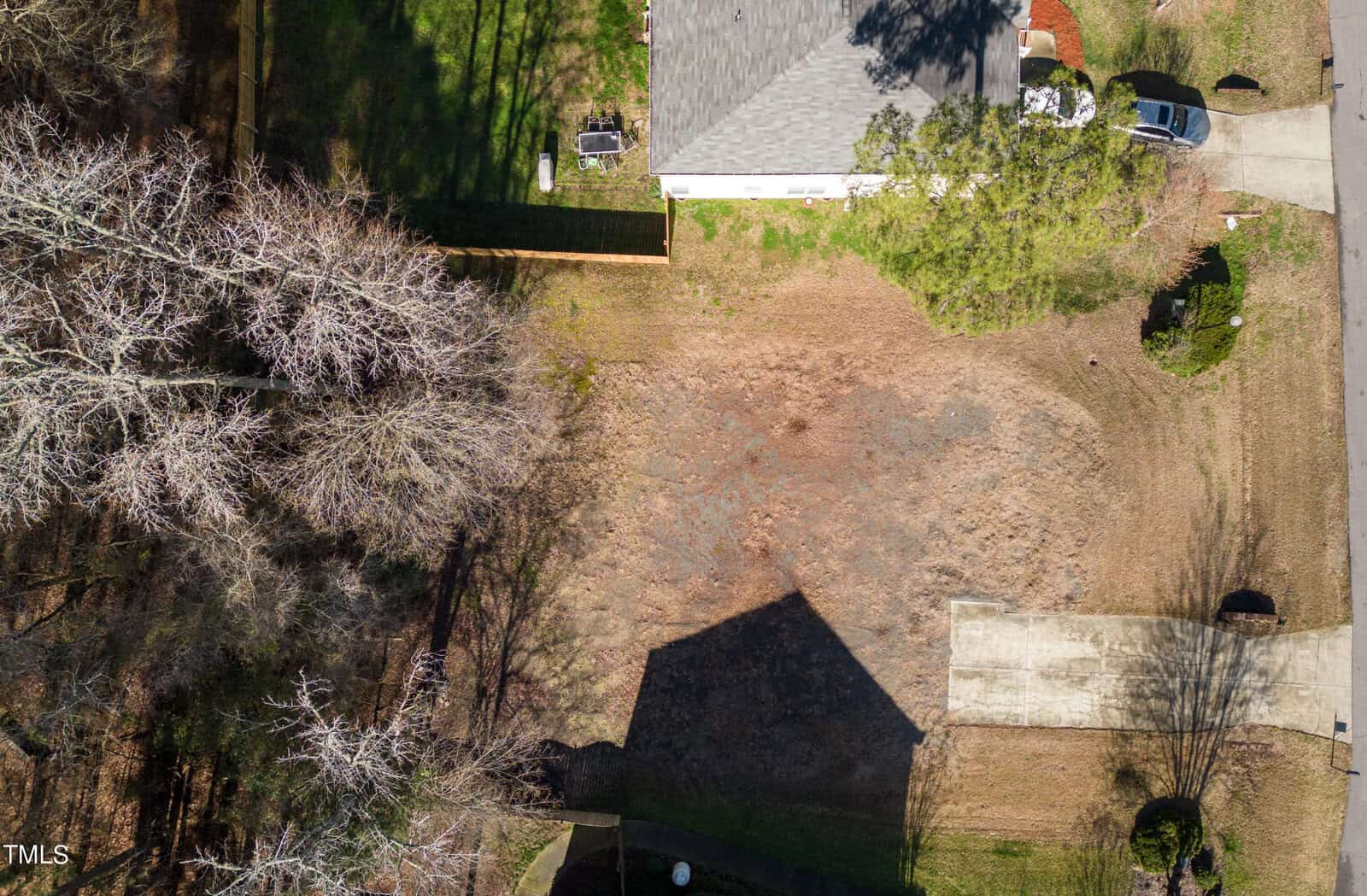 Aerial view of a residential area with a single house and driveway on the right. The center of the image shows a large, empty plot of land with sparse grass. Bare trees and shadows are visible on the left and in the foreground.