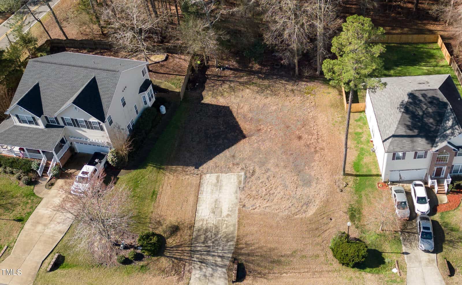 Aerial view of an empty residential lot with a driveway, flanked by two houses. The lot is covered in brown grass, and trees are visible in the background. Each house has a driveway with parked vehicles.