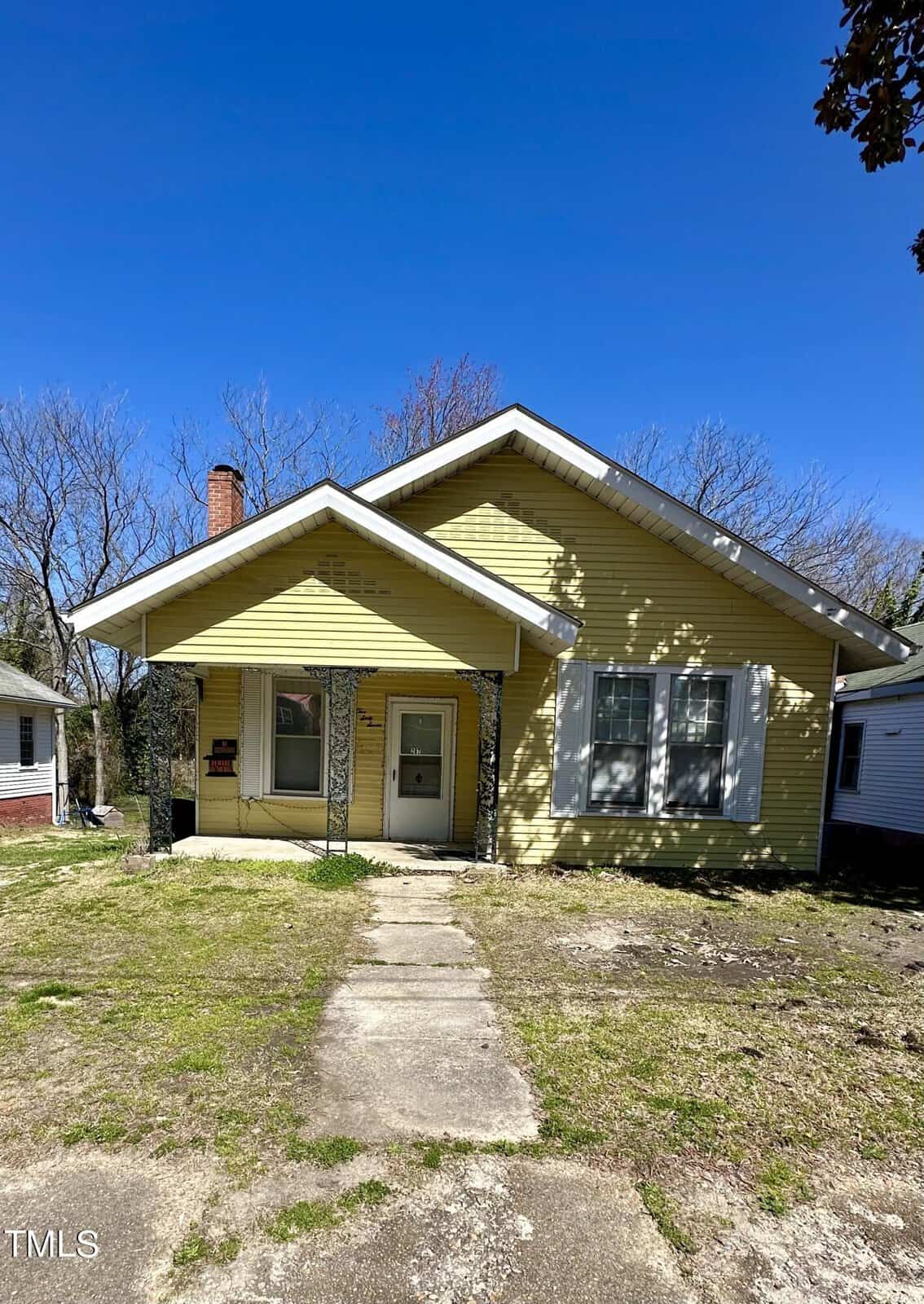A single-story yellow house with a gabled roof and white trim, featuring a front door with a sidelight and concrete walkway leading to the entrance; a chimney is visible on the left side of the house.