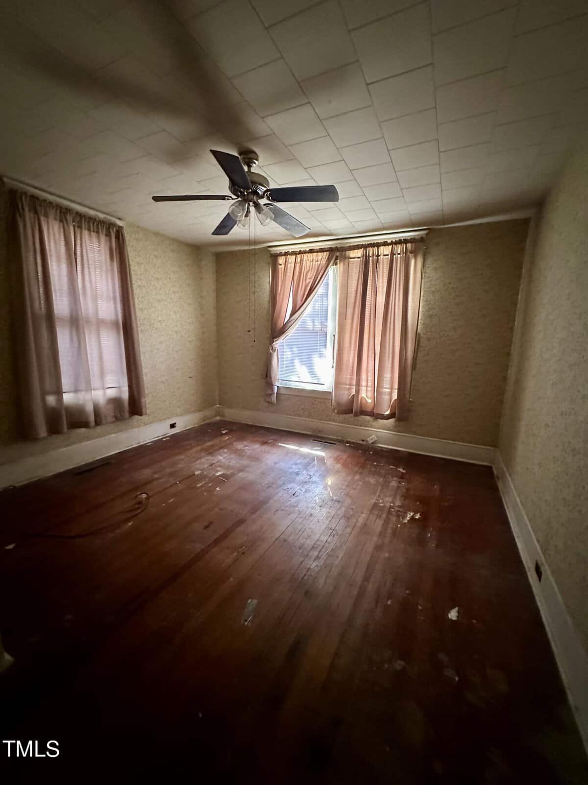 An old, empty room with worn hardwood floors, a ceiling fan, and aged, partially drawn curtains on a window letting in dim light.