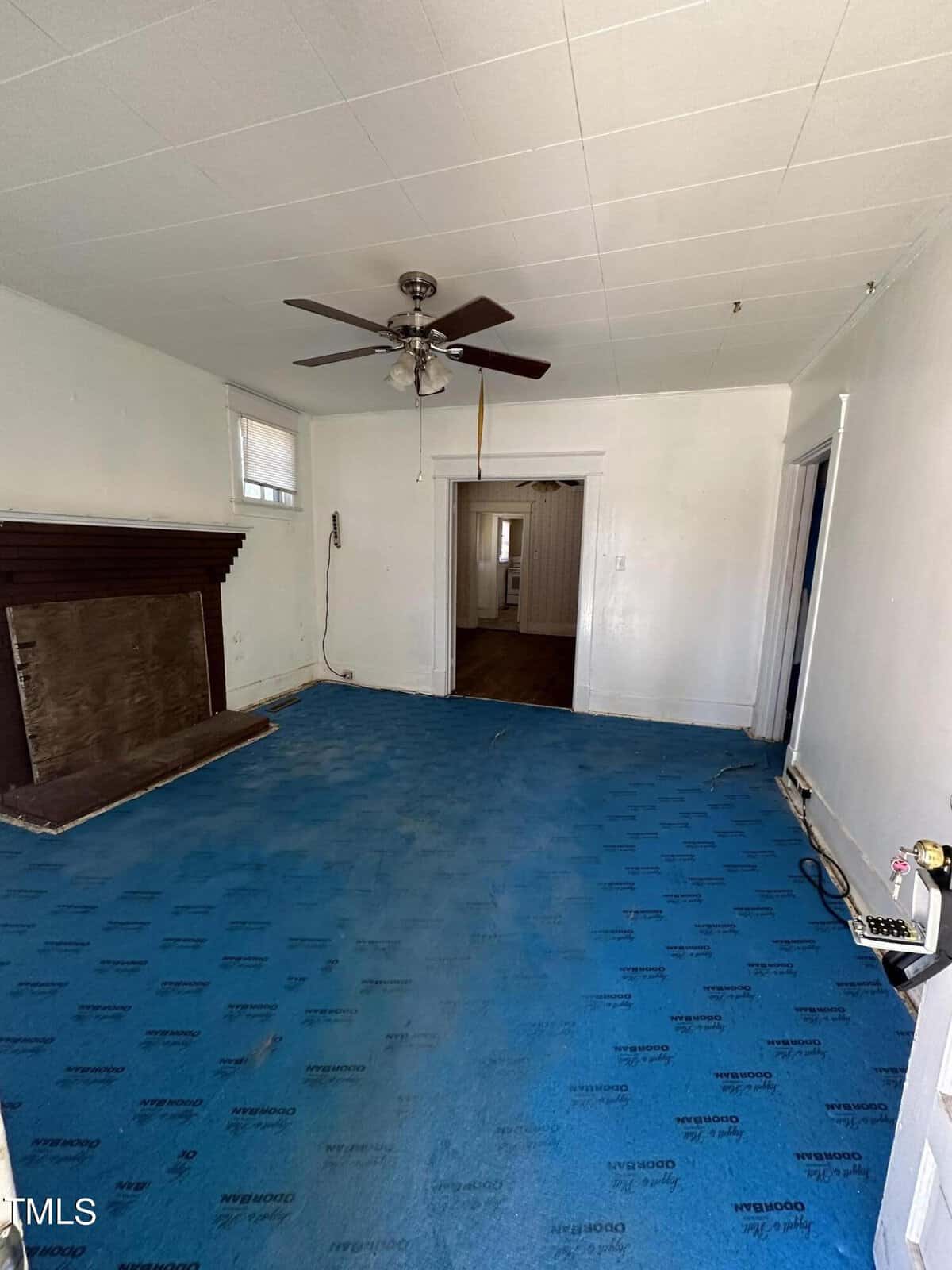 An empty room featuring a blue carpet with a repetitive design, white walls, a ceiling fan, and a fireplace on the left side. the view through an open door shows another room in the background.