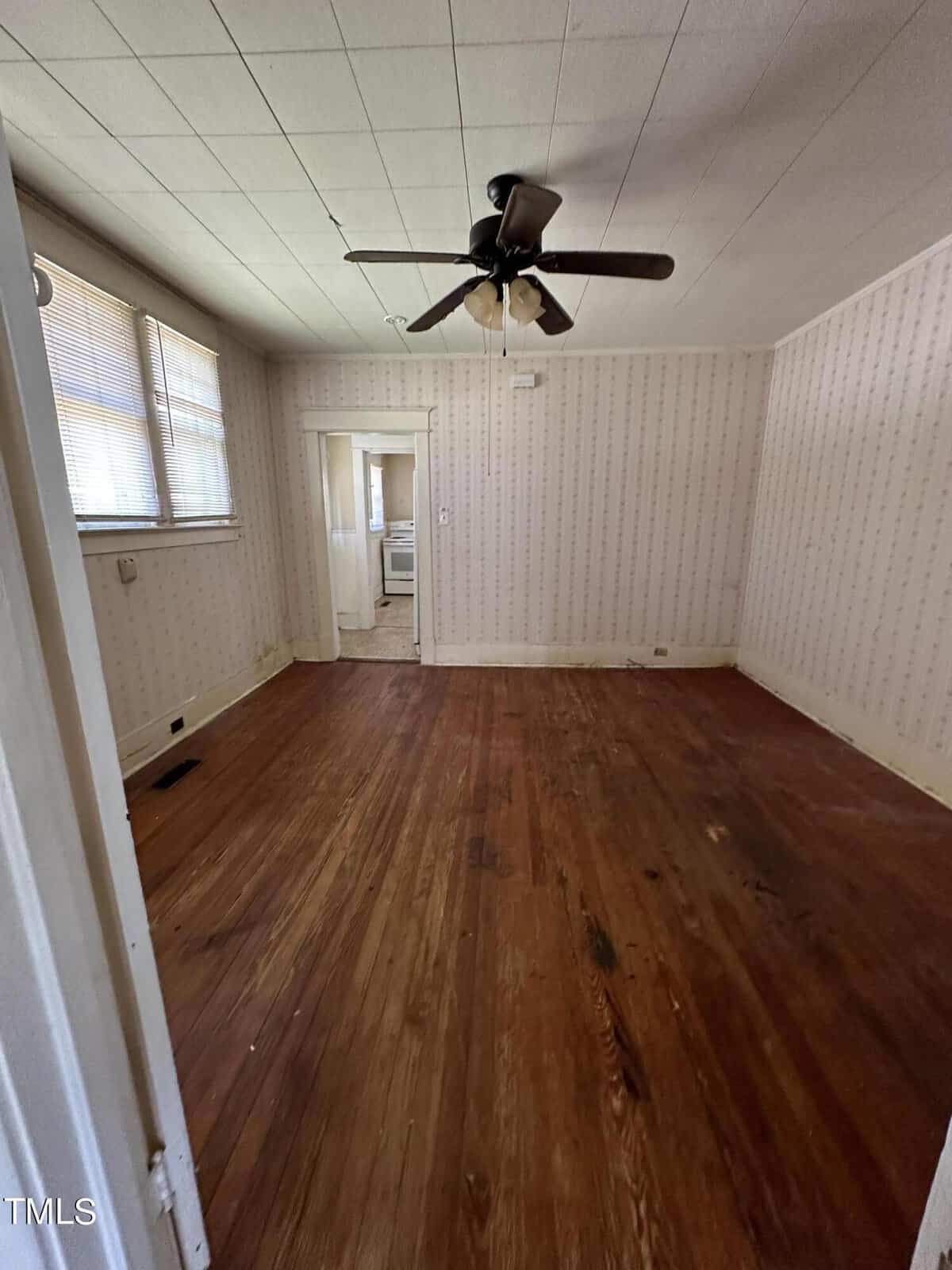 An empty room with wood flooring, patterned wallpaper, a ceiling fan, and a door leading to another room.