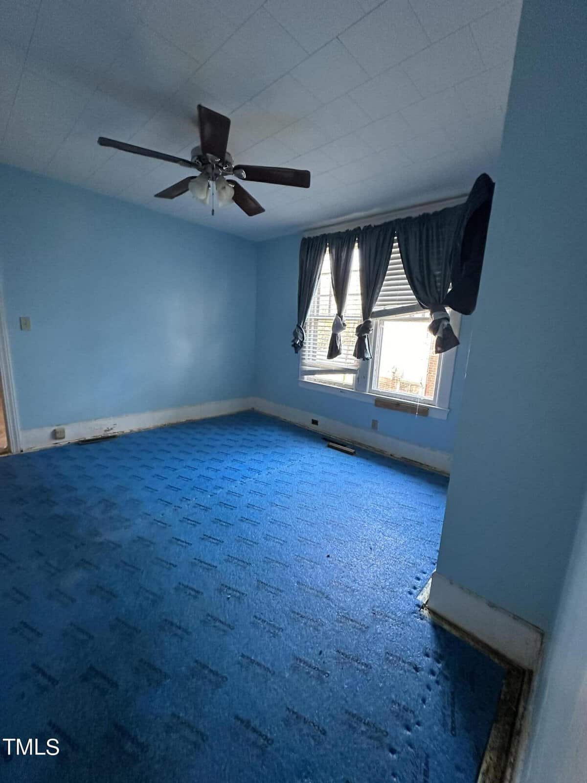 A vacant room with blue walls, matching blue carpet, a ceiling fan, and draped windows allowing natural light to enter.