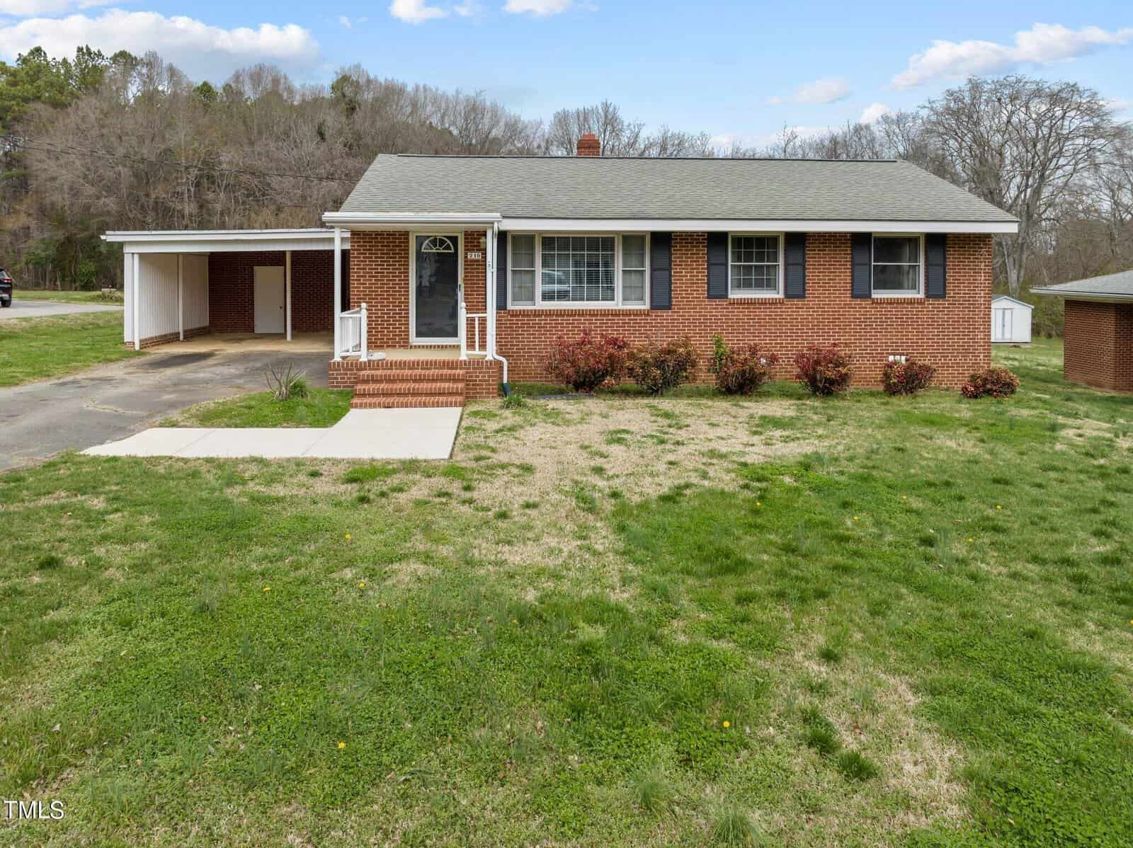Brick house with a gray roof, white-framed windows, and a covered carport on the left. Bushes line the front of the house. The yard is grassy with some patches. Trees and a blue sky with clouds are in the background.