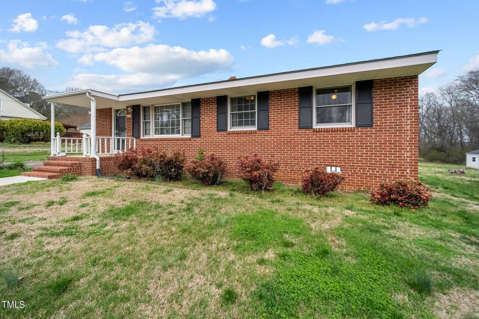 A single-story red brick house with a white front porch. The house has black shutters and a sloped grassy yard with shrubs. The sky is partly cloudy.