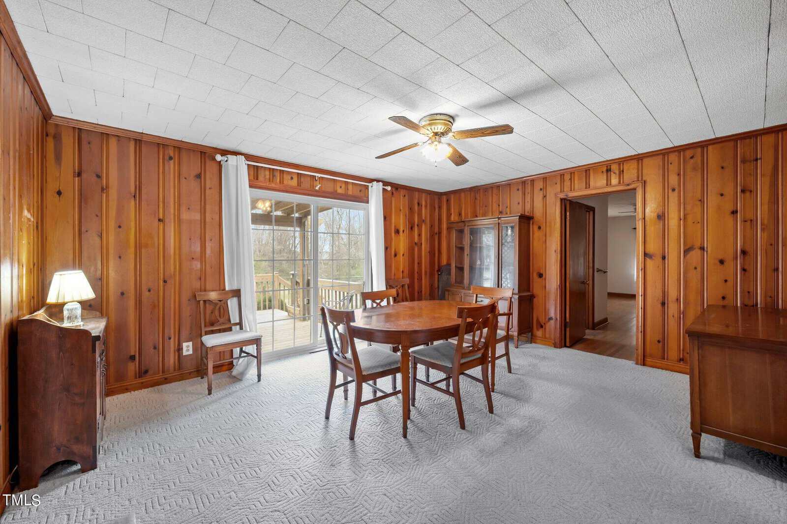A wood-paneled dining room with a round wooden table and four chairs. There's a cabinet against the wall, a door to the right, and sliding glass doors leading to an outdoor deck. A ceiling fan with lights is centered above.