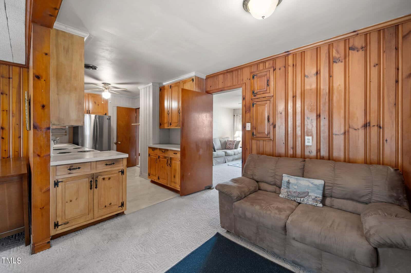 Living room with wood-paneled walls and a brown sofa. The room leads to a kitchen with wooden cabinets, white countertops, and stainless steel appliances. A glimpse of a carpeted room with a sofa is visible in the background.