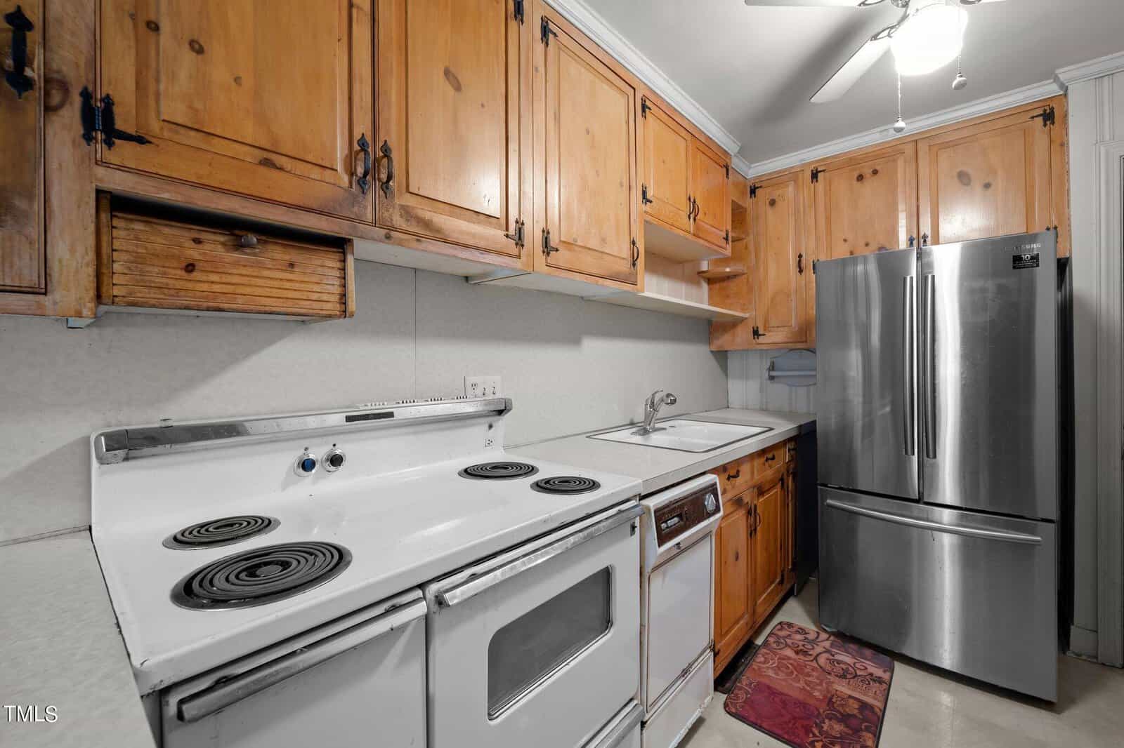 A kitchen with wooden cabinets, a vintage white electric stove, a dishwasher, and a modern stainless steel refrigerator. The room has a white ceiling with a light and fan. A small rug lies on the floor near the sink.