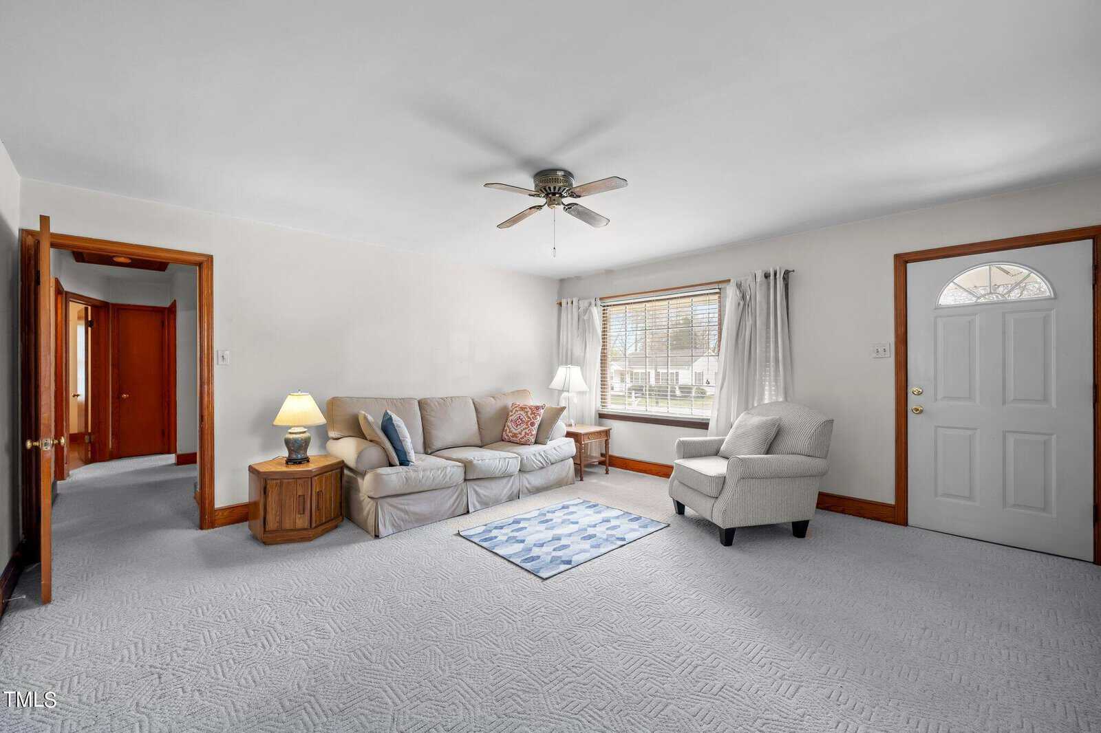 A cozy living room with a beige sofa, patterned armchair, and wooden side tables with lamps. A ceiling fan is mounted above and a window with white curtains lets in light. The floor features a blue patterned rug. Doorways lead to other rooms.