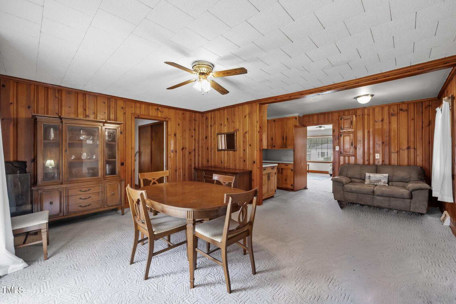 A mid-century style dining room with wood-paneled walls and carpeted floor. It features a ceiling fan, a wooden dining table with four chairs, a china cabinet, and a view into the kitchen and living area with a couch.