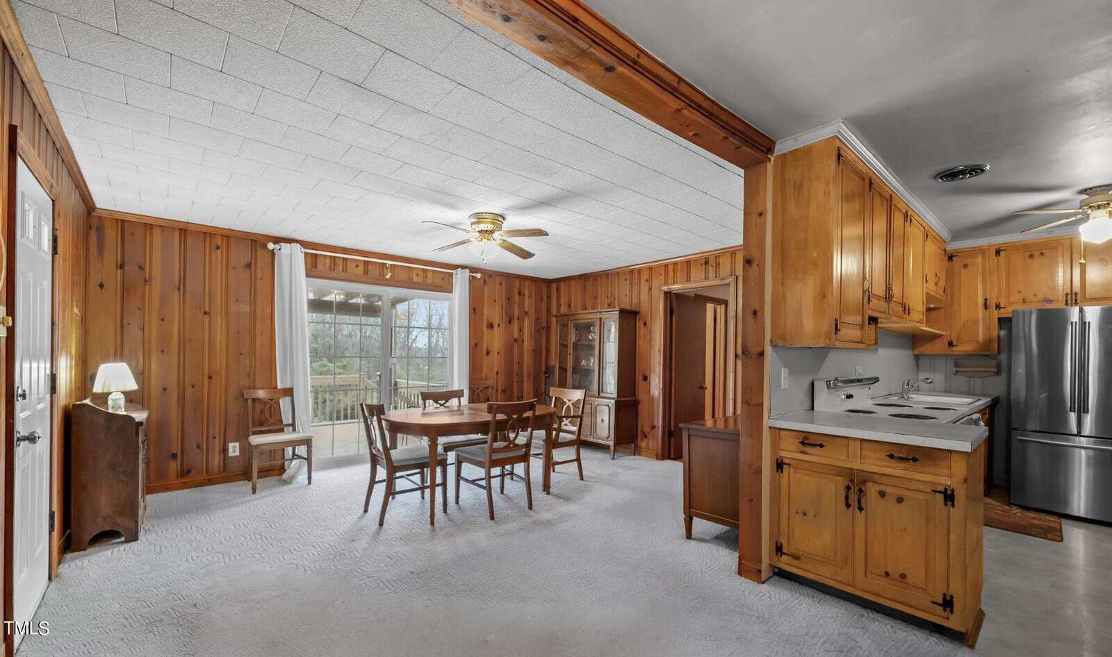 A cozy dining room with wood-paneled walls, carpeted floor, and ceiling fans. It features a wooden dining table with chairs, a cabinet, and a view of a balcony with trees outside. The adjacent kitchen includes wood cabinets and stainless steel appliances.
