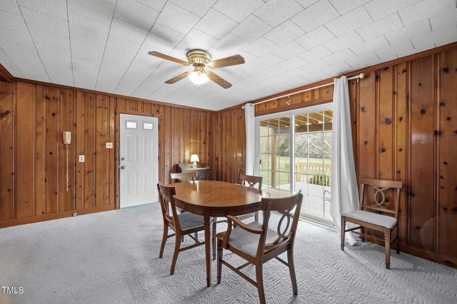 Dining area with wooden panel walls and a ceiling fan. A round wooden table is surrounded by chairs. There is a glass sliding door leading to a patio, letting in natural light, and a small desk with a lamp in the corner.